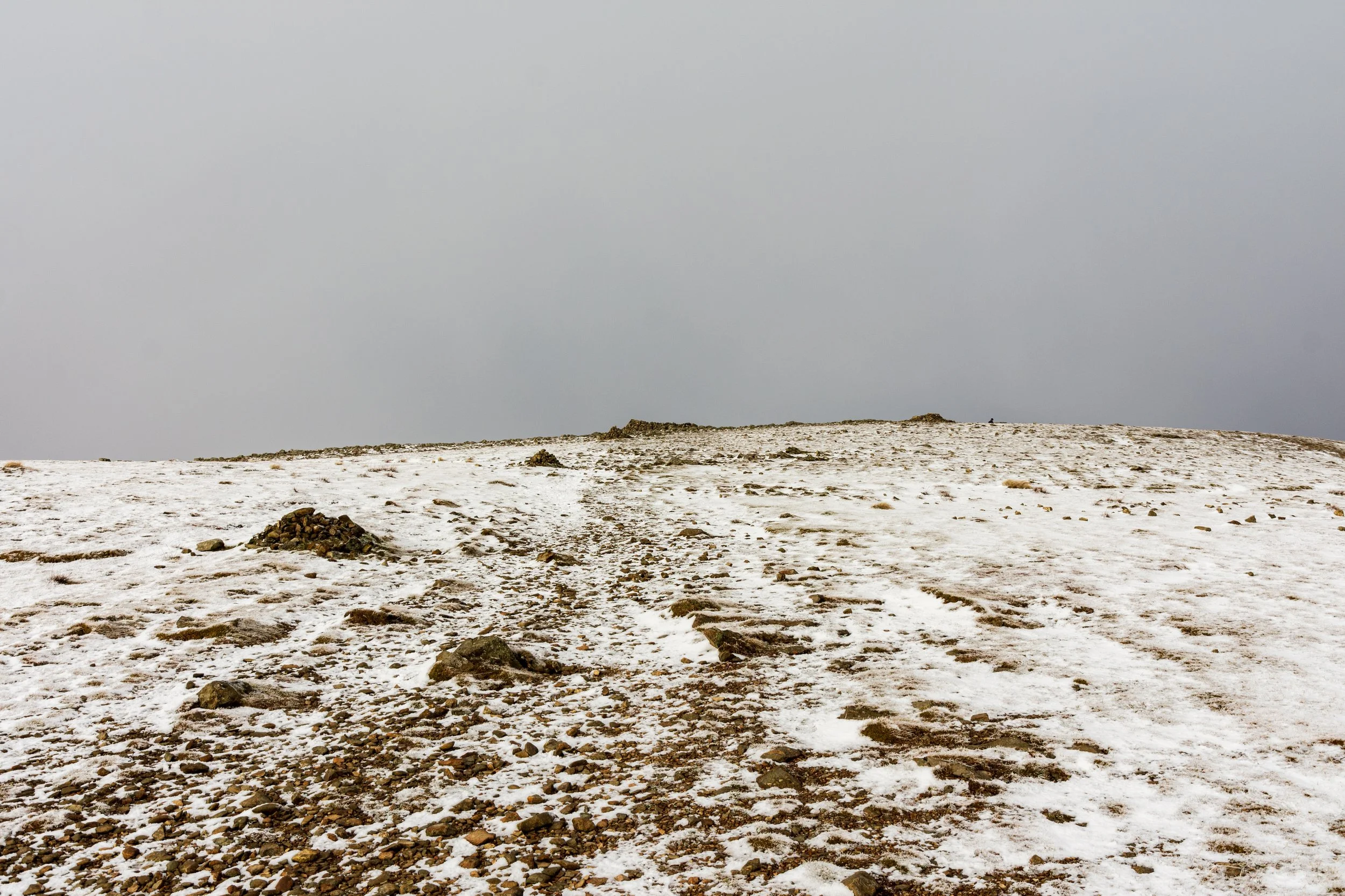 A snowy summit of Fairfield in the Lake District, showing cairns and a shelter, amidst a cloudy grey sky.