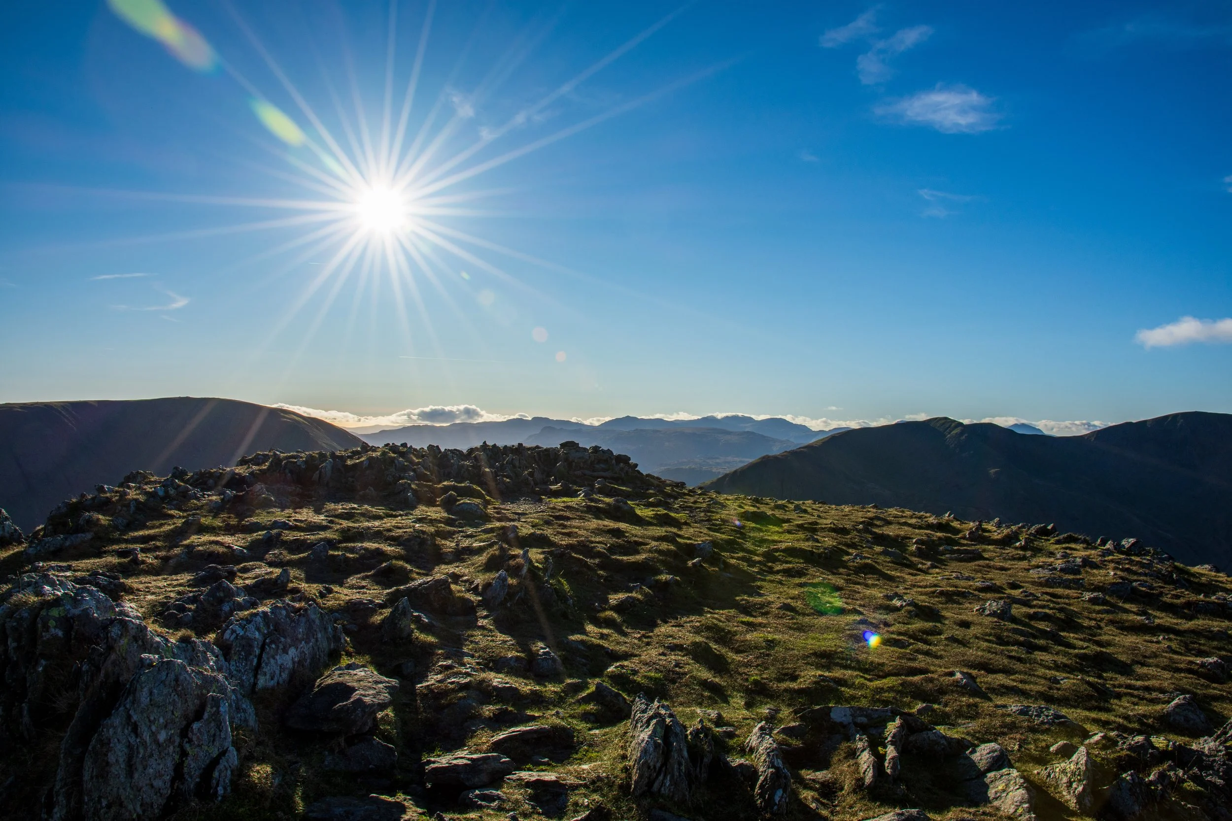 A view from the summit of St Sunday Crag in the Lake District towards the west, with Fairfield on the left and Dollywaggon Pike on the right. The summit is rocky, the sky is blue, with the later afternoon sun in bright in the sky