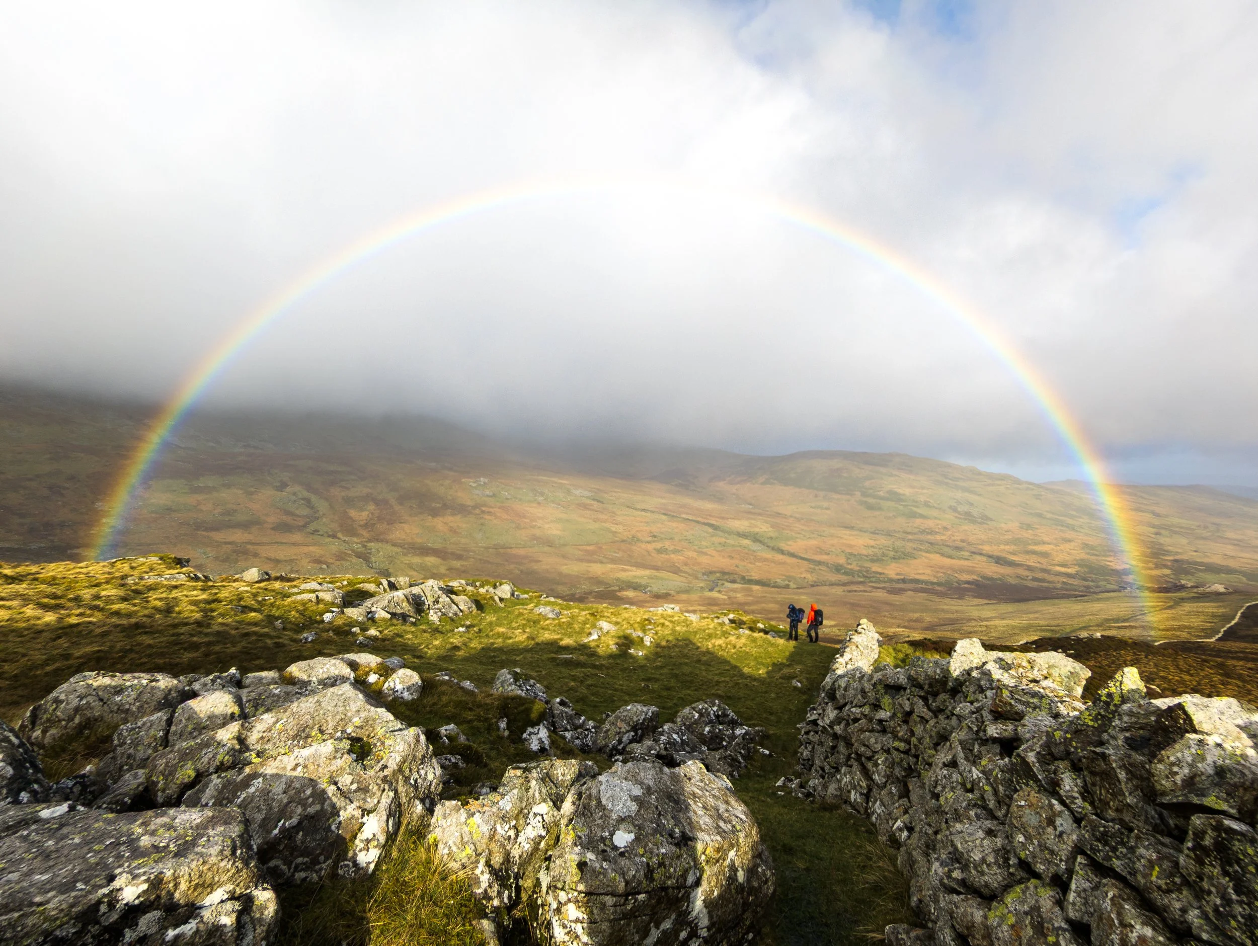 Two people on a 1:1 Business Coaching session in the mountains of Snowdonia, with a rainbow across a stormy sky, and rocks in the foreground