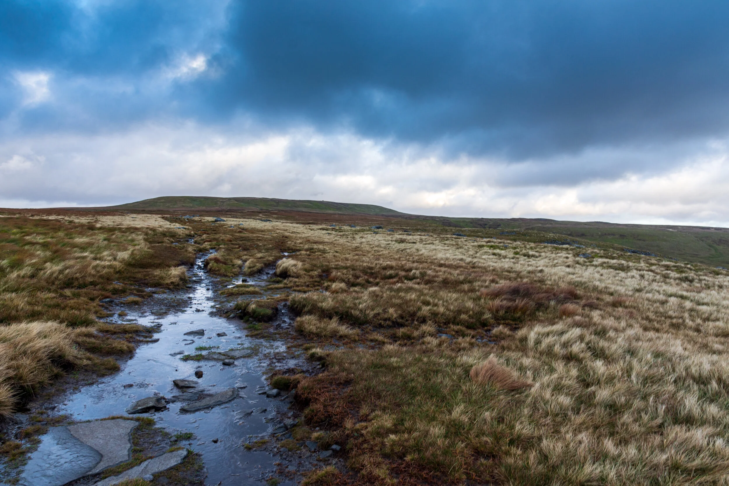 A boggy, peat covered path amongst the remote moorland approaching the summit of Great Shunner Fell, against a dramatic, sky