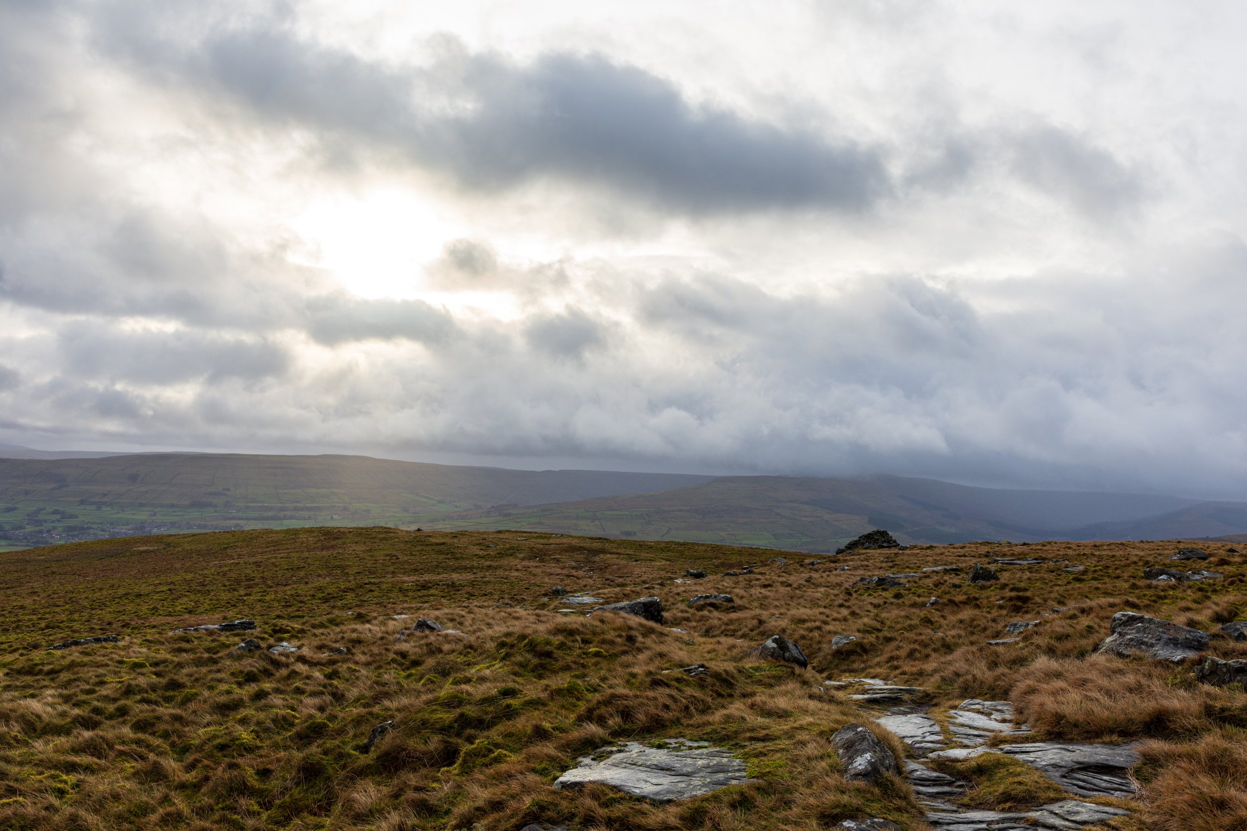 A wet, boggy moor with rocky outcrops and a cairn on the lower slopes of Great Shunner Fell, looking over Wenselydale and into the Yorkshire Dales. A stormy sky with low cloud, and the sun breaking through
