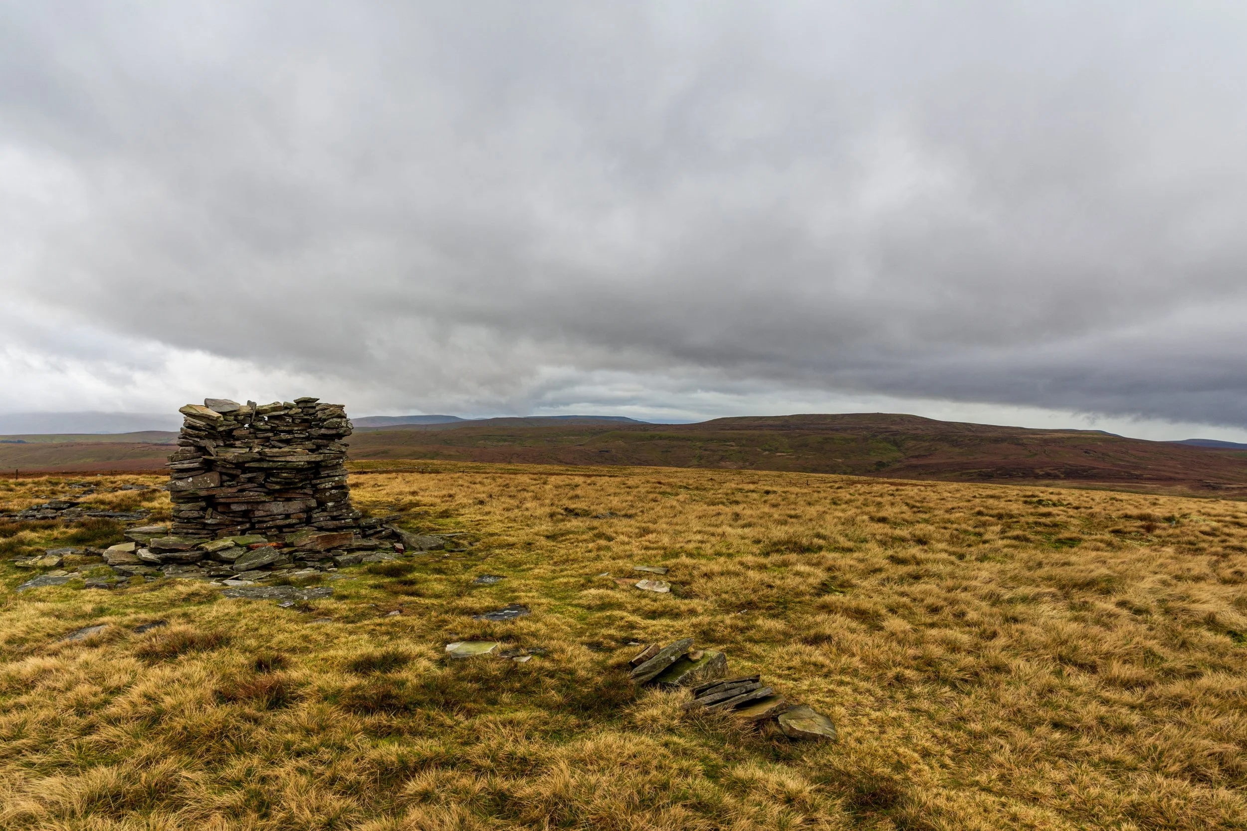 Ancient cairn on the summit of Lovely Seat on desolate moorland, during a walk in the Yorkshire Dales, against a stormy sky and Swaledale in the background