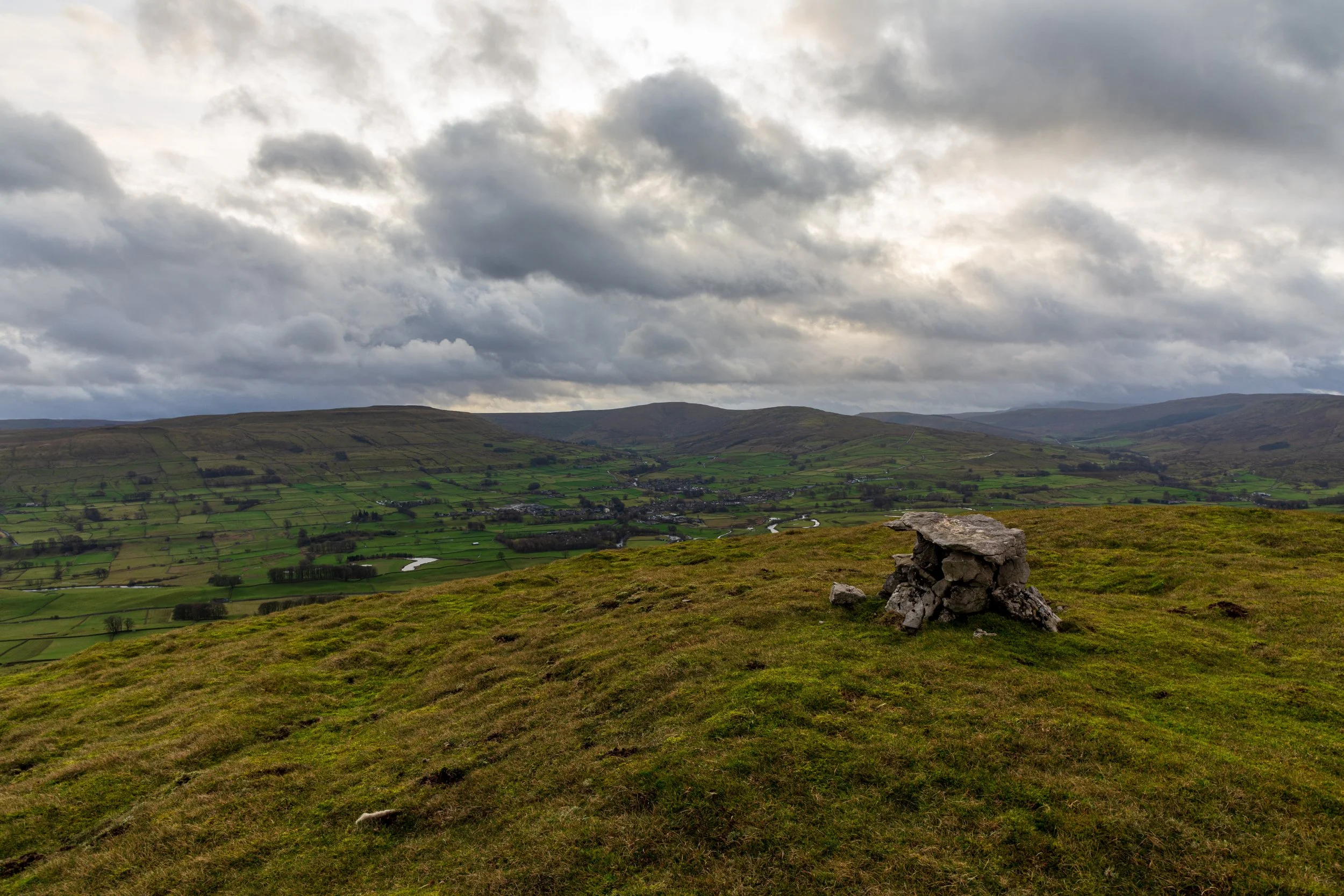 A view of the cairn on the summit of Smuker Hill, on the lower slops of Lovely Seat, overlooking Wenseleydale, on a walk in the Yorkshire Dales. Sun is breaking through the stormy sky, and the valley is green and wet