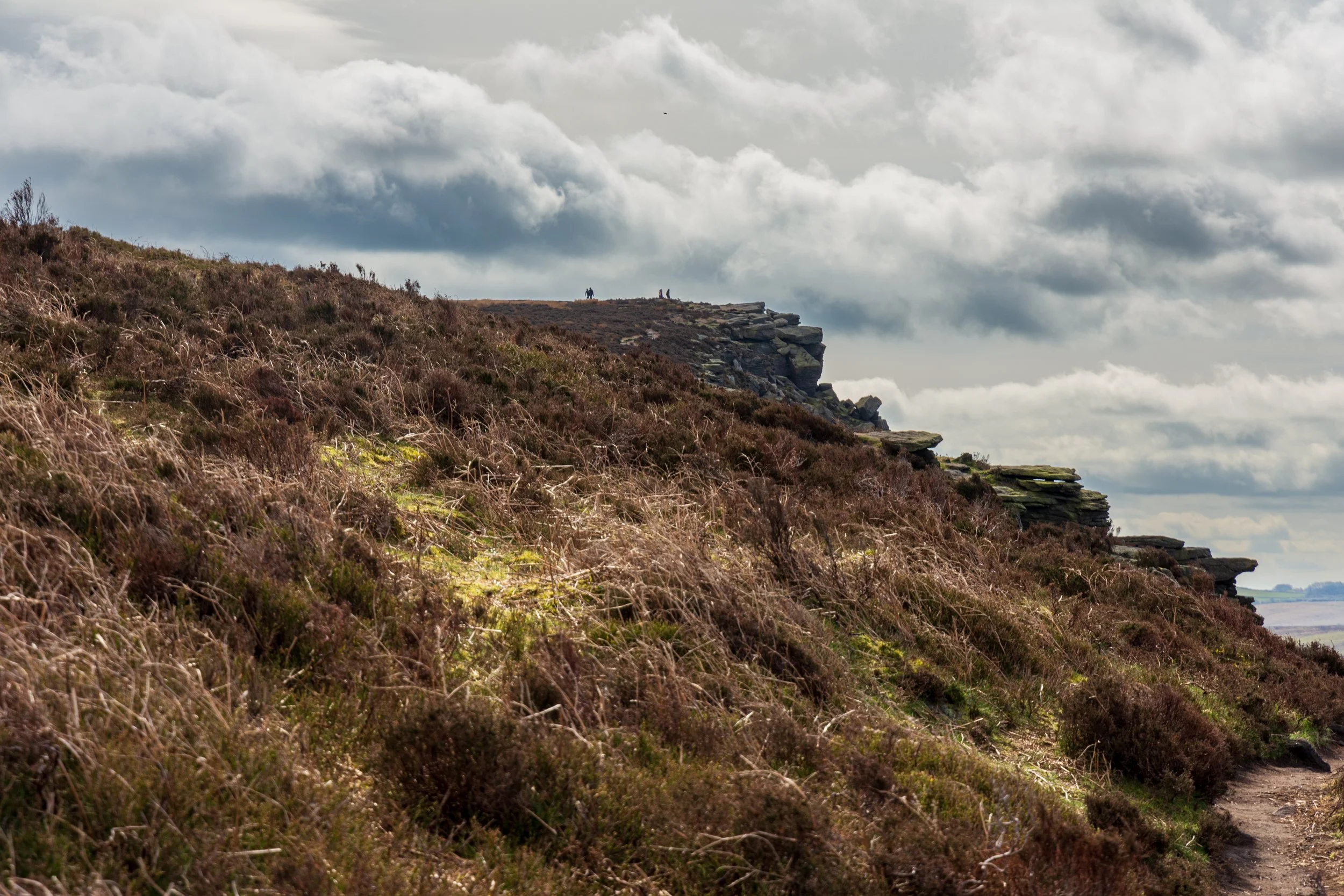 A view of Great Tor on Bamford Edge in the Peak District from a heather moor below and to the North, with silhouettes of walkers on the top, under a cloudy sky