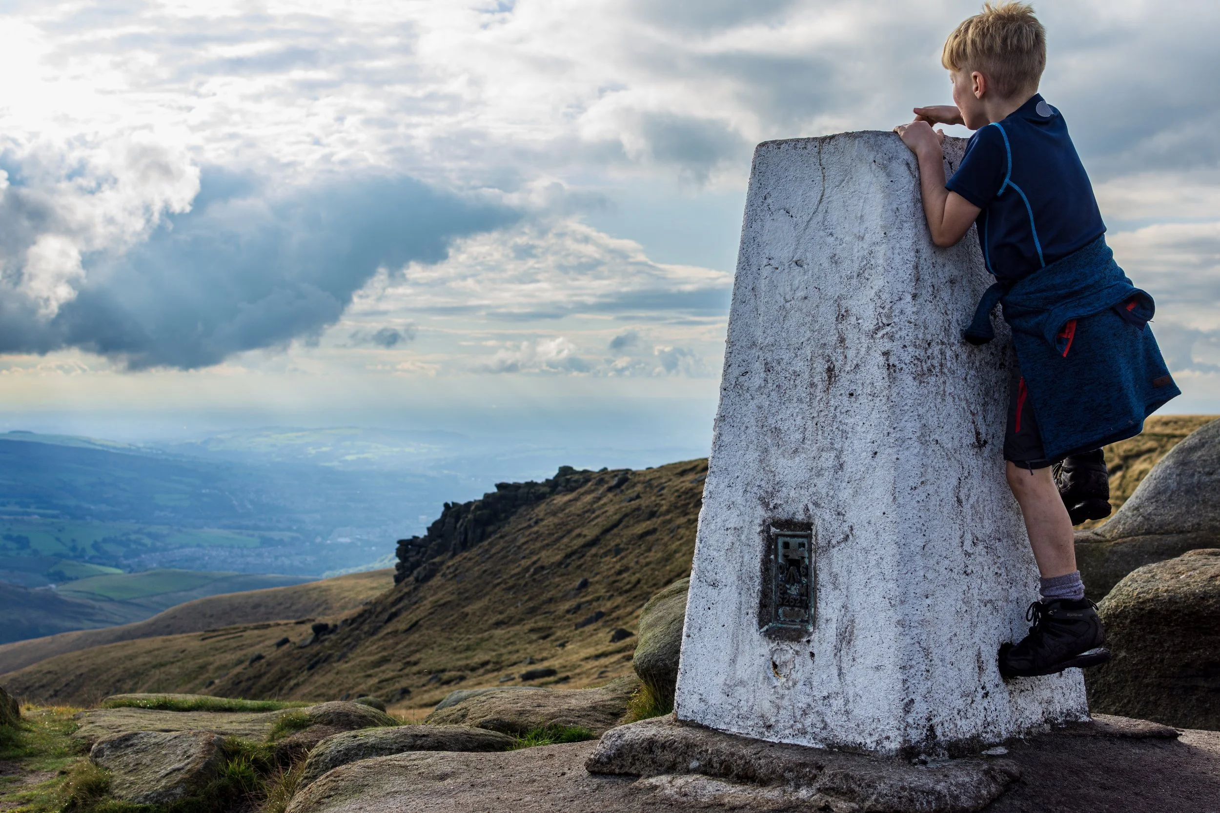 7 year old boy climbing the white trig pillar at Higher Shelf Stones, Peak District, with a clear view overlooking the valley and Manchester