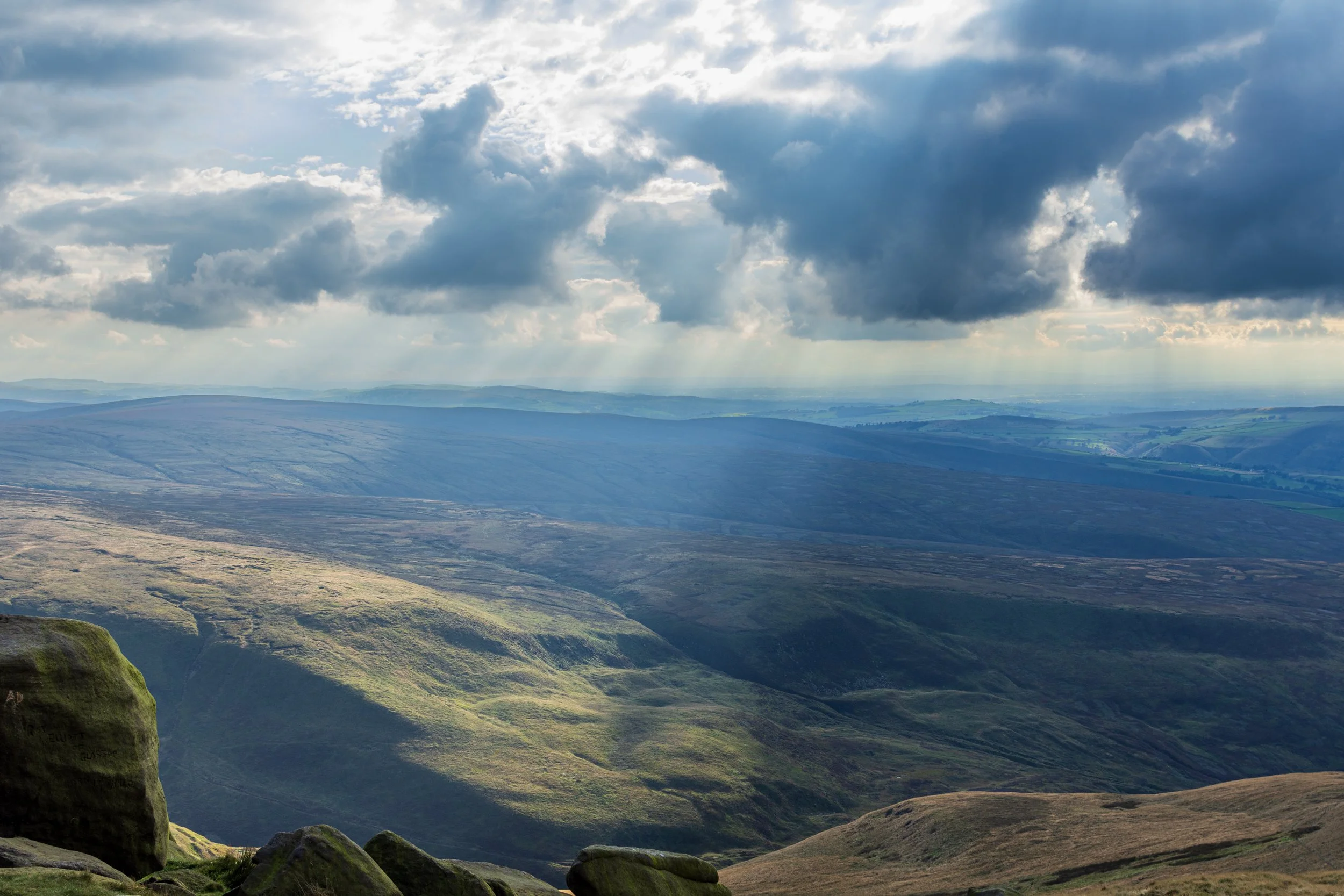 View of the rugged and rolling hills of the Peak District, from Higher Shelf Stones, against a cloudy sky, with cloudbursts shining sunlight onto the landscape