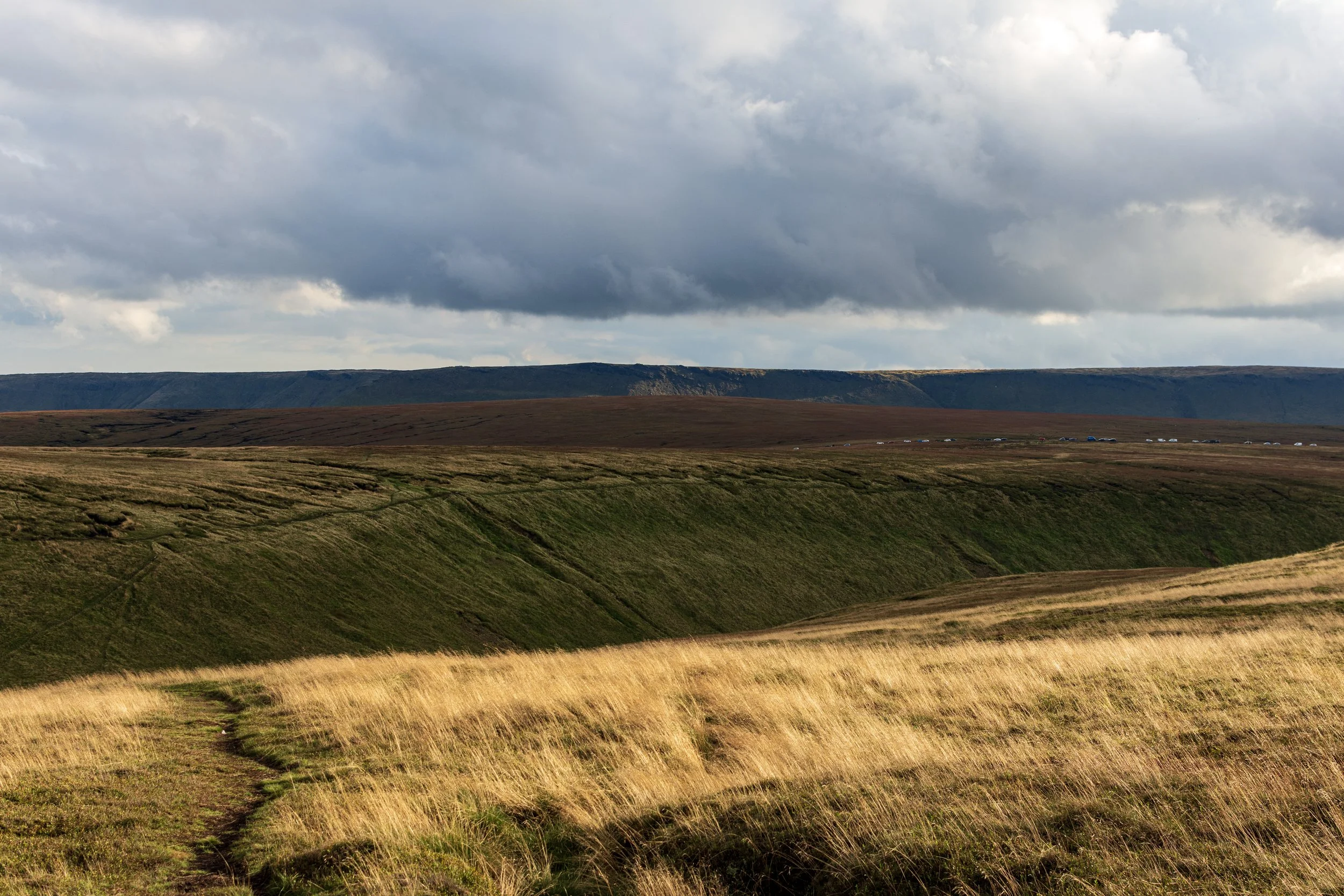 View of the moors below Higher Shelf Stones in the Peak District, looking at the deep cut into the moor which is Crooked Clough. The Snake Pass summit is beyond, with cars parked on the road. In the background is Kinder Scout, lit by the sun