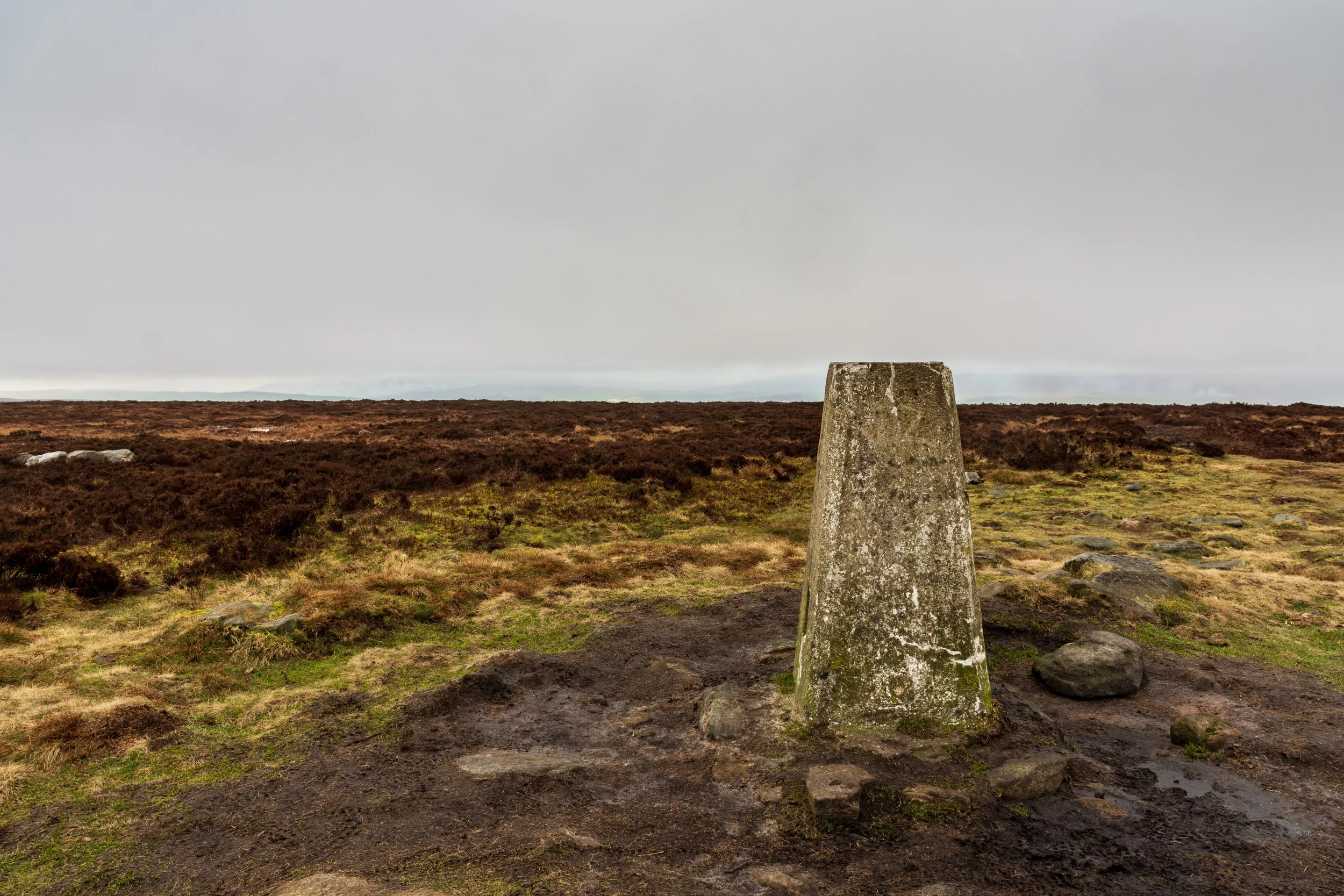 The weathered trig point on the summit of Ilkley Moor / Rombalds Moor, on an open heather moorland, with a rain-filled sky