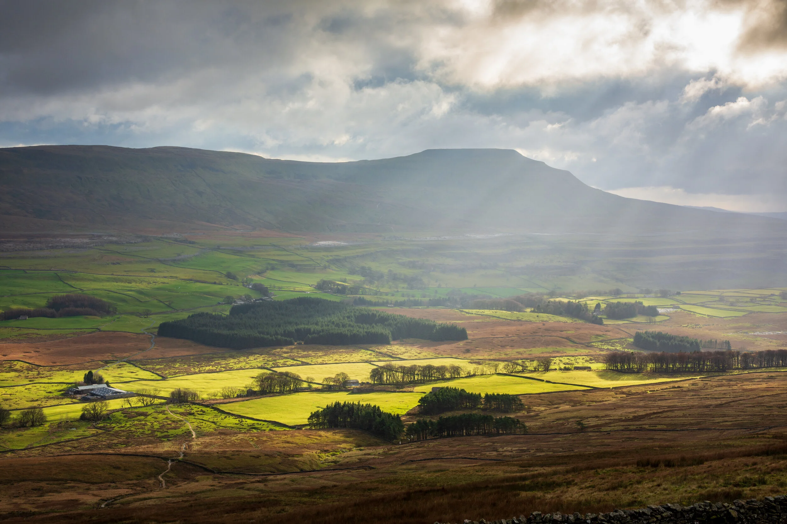 Looking at Ingleborough, across the Ribble Valley in the Yorkshire Dales, from the slopes of Whernside. The valley is green with fields and trees, sunlit by a cloud burst from a stormy sky.