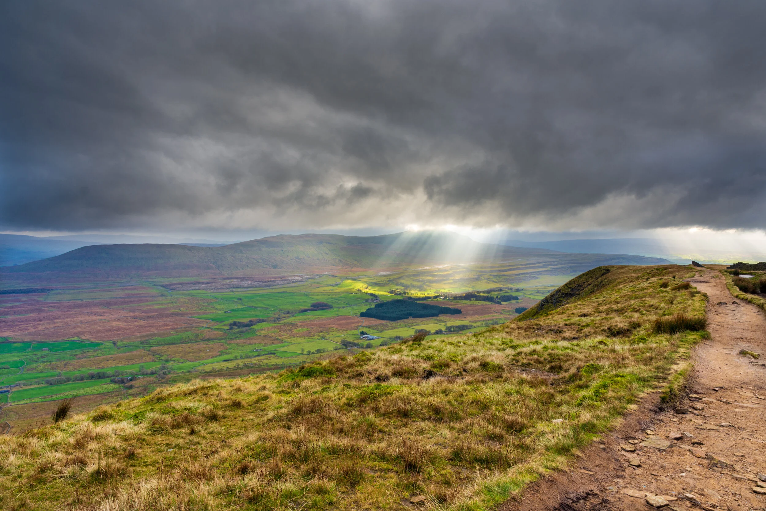A path near the summit of Ingleborough, looking across the green Ribblesdale in the Yorkshire Dales, a very grey stormy sky with a cloudburst lighting up a small wood. Whernside is in the background