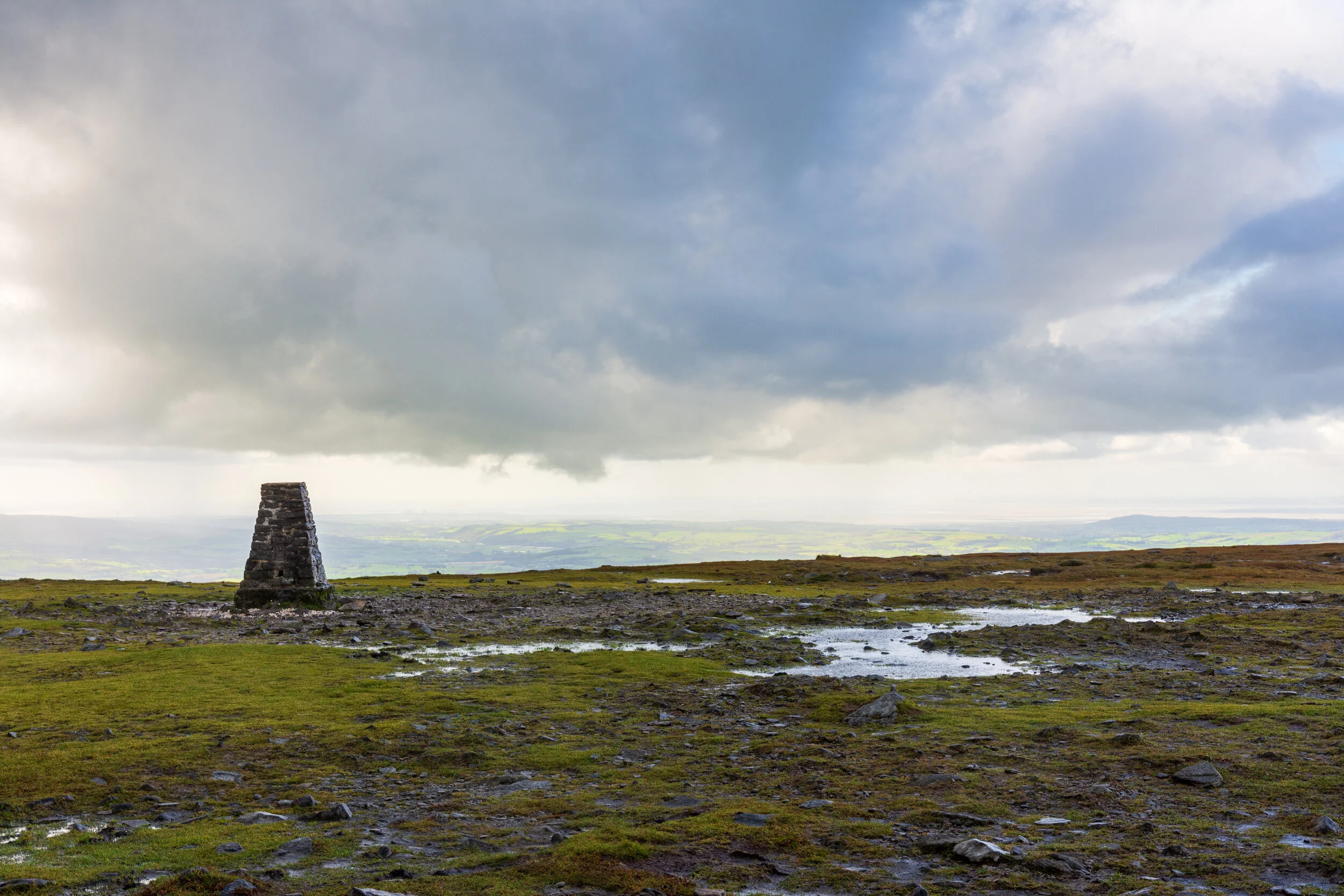 The flat summit of Ingleborough, covered with grass and broken rocks. The trig pillar is in the mid-ground, and the view looks out towards Lancashire and Morecambe Bay. The sky is cloudy but bright and the ground is wet