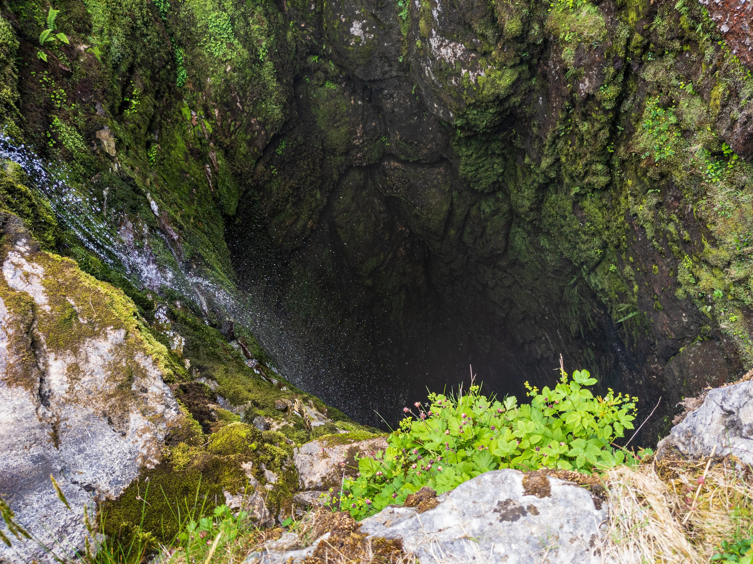 A view from the top of Gaping Gill on the slopes of Ingleborough in the Yorkshire Dales, looking down into the pothole. The moss covered rock walls disappear into the darkness, and spray from the waterfall hitting the sides can be seen