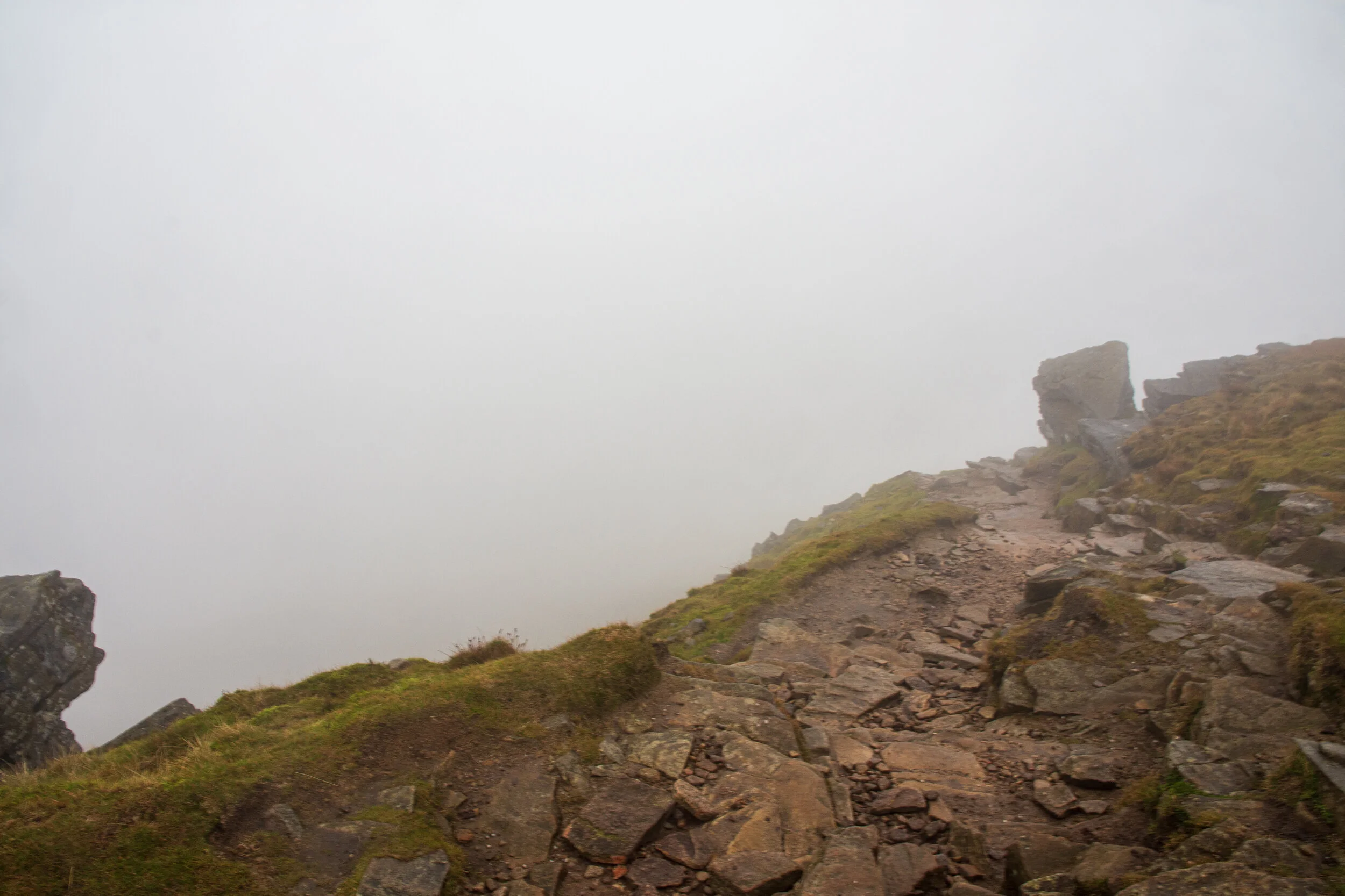A rocky path on the summit of Ingleborough. The ground falls away steeply to the left into the foggy abyss. There is no view because of the clouds and mist