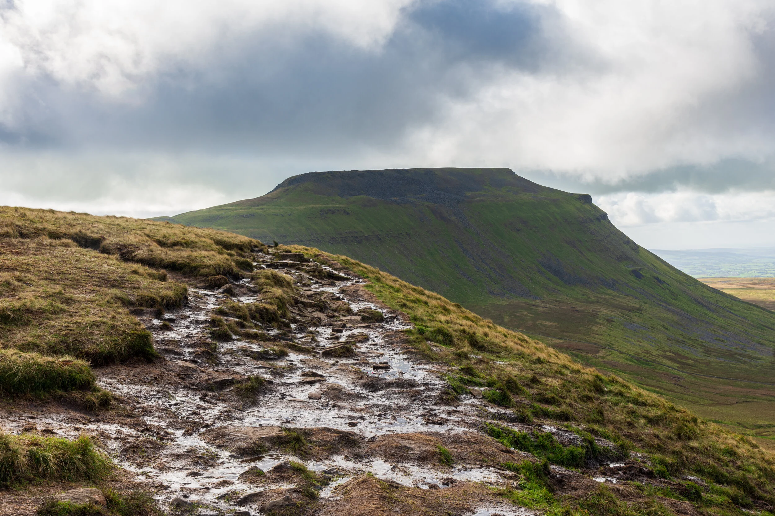 A muddy and wet path between Simon Fell and Ingleborough in the Yorkshire Dales. The ground drops steeply to the right into Ribblesdale, the green slopes of Ingleborough and its flat summit are prominent in the mid-ground. Cloudy sky, brightening up