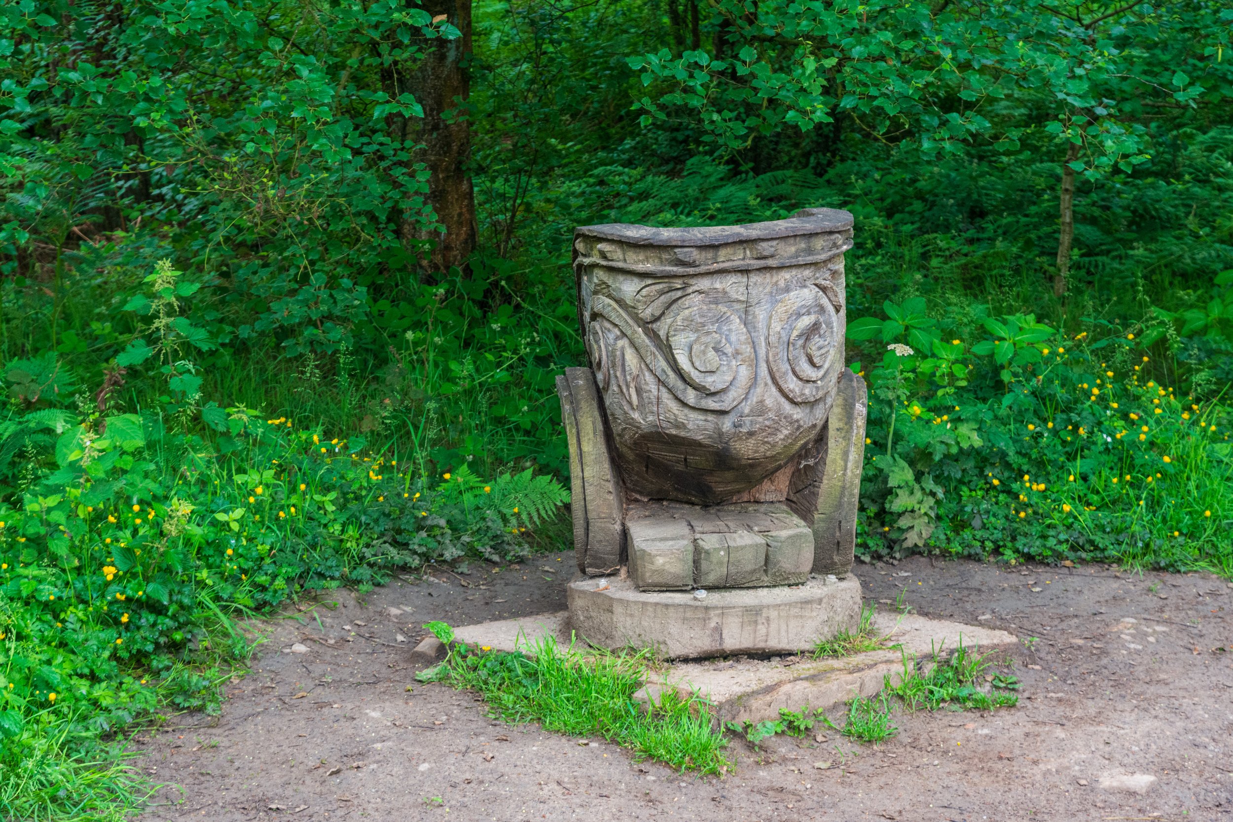 A wooden cauldron sculpture, decorated with an ornate pattern, in a clearing amongst trees and greenery in Otley Chevin, Yorkshire