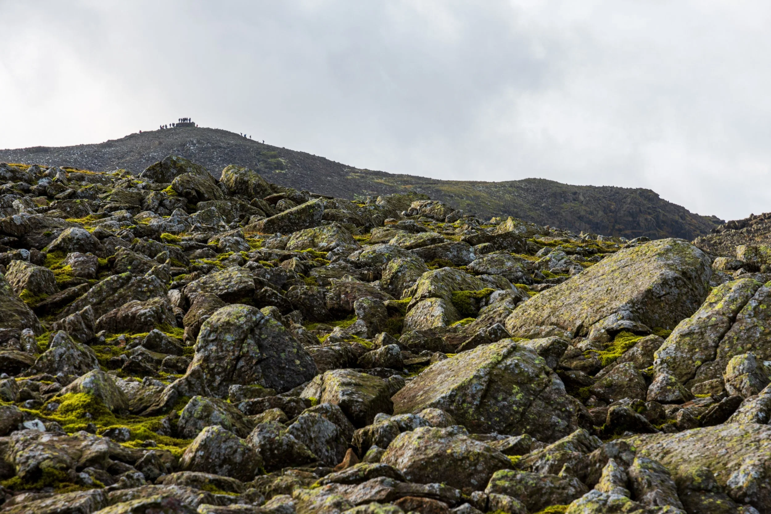The grey-rock and moss-strewn boulder field of Esk Pike in the Lake District in the foreground, with Scafell Pike in the background, with approx. 20 people visible on the summit and standing on the shelter. Cloudy day with some sunshine
