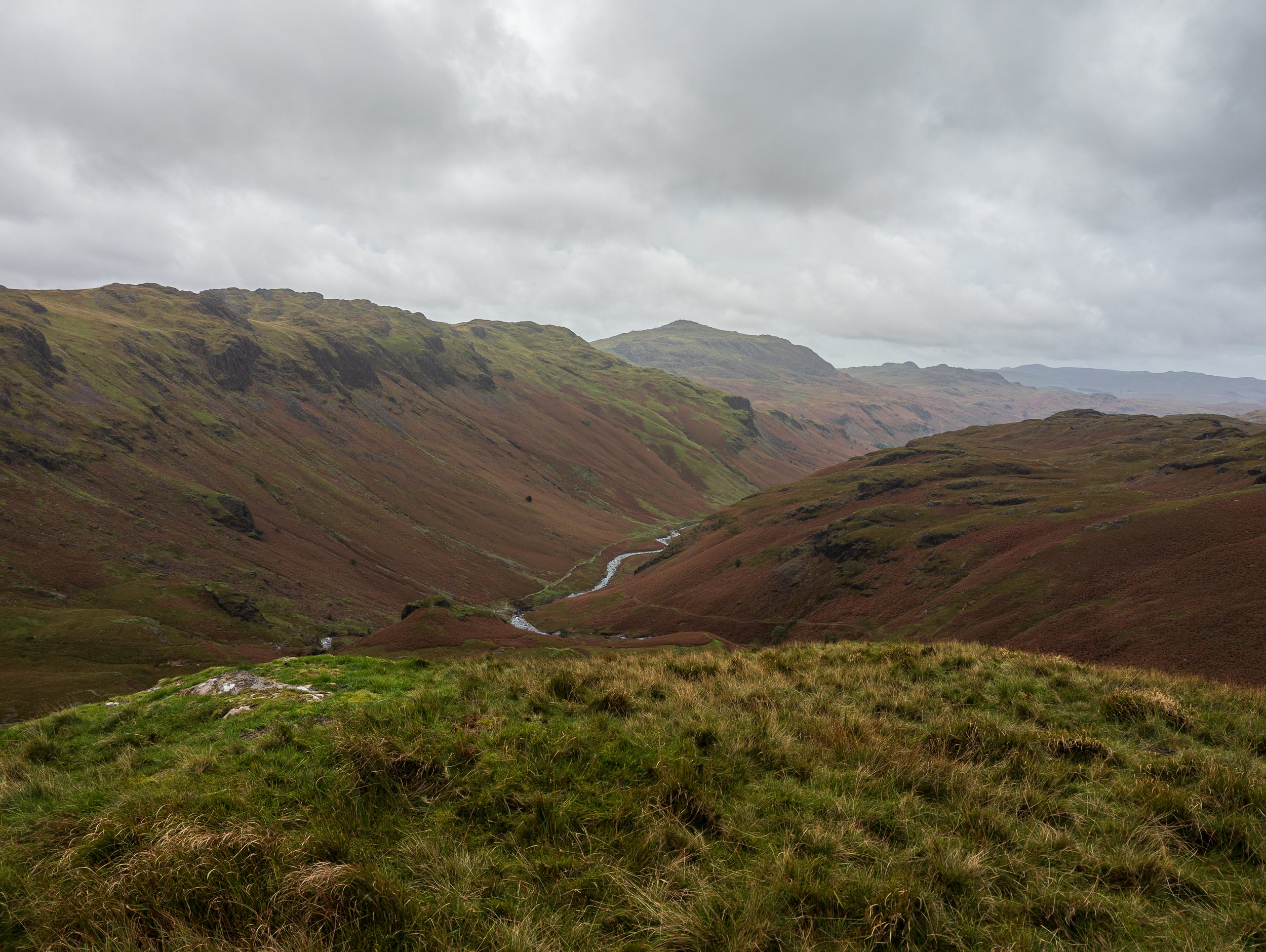 A view of a stream running through the valley of Eskdale, taken from the top of Throstlehow Crag, with Cam Spot Crag, Scafell Pike and Ill Crag visible, overcast day with barren brown moors