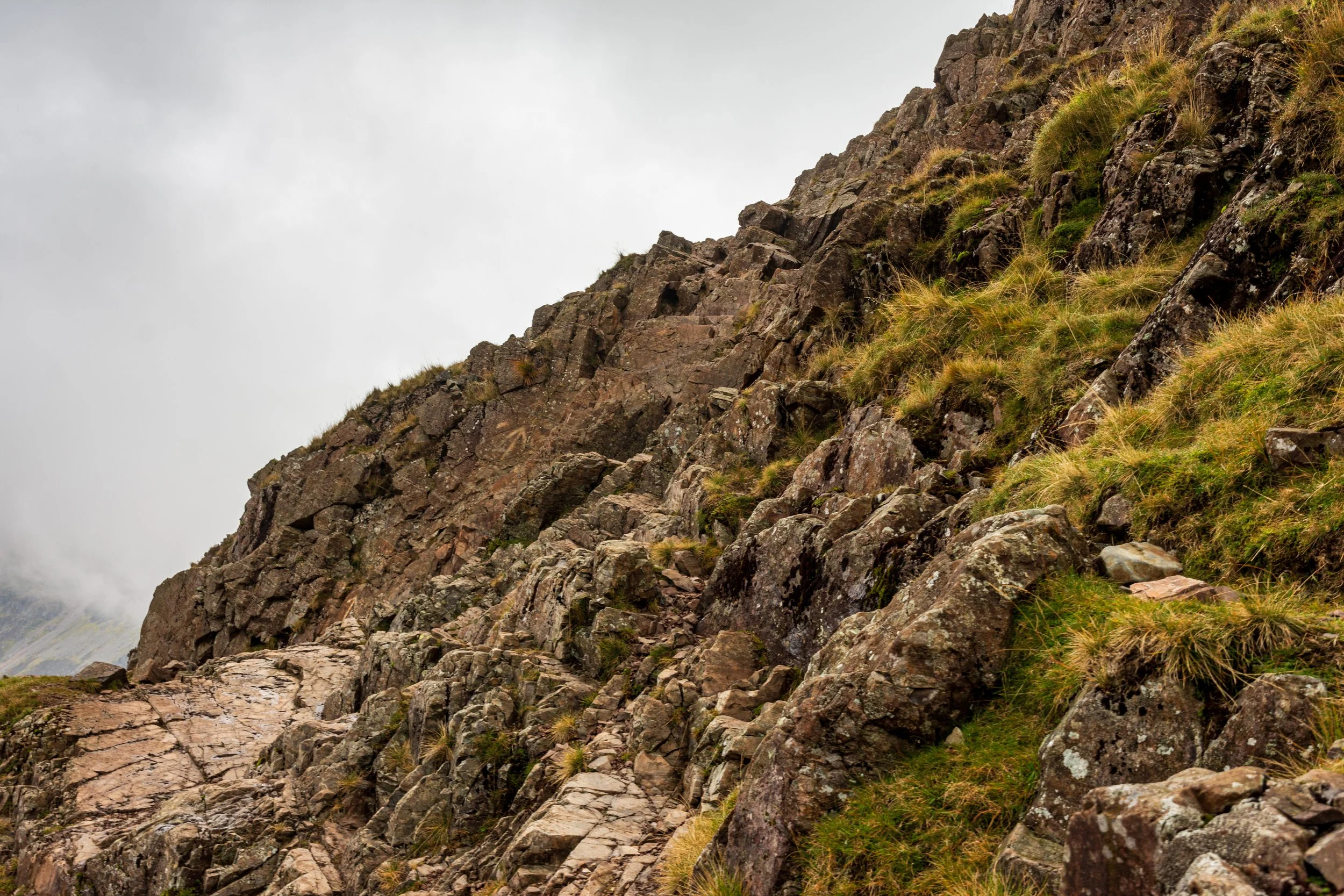 A rocky bad step on the Corridor Route on the way up to Scafell Pike in the Lake District. A series of rock steps in the side of the mountain, surrounded by a steep drop to one side, and a grassy area to the other. Summits enveloped in cloud