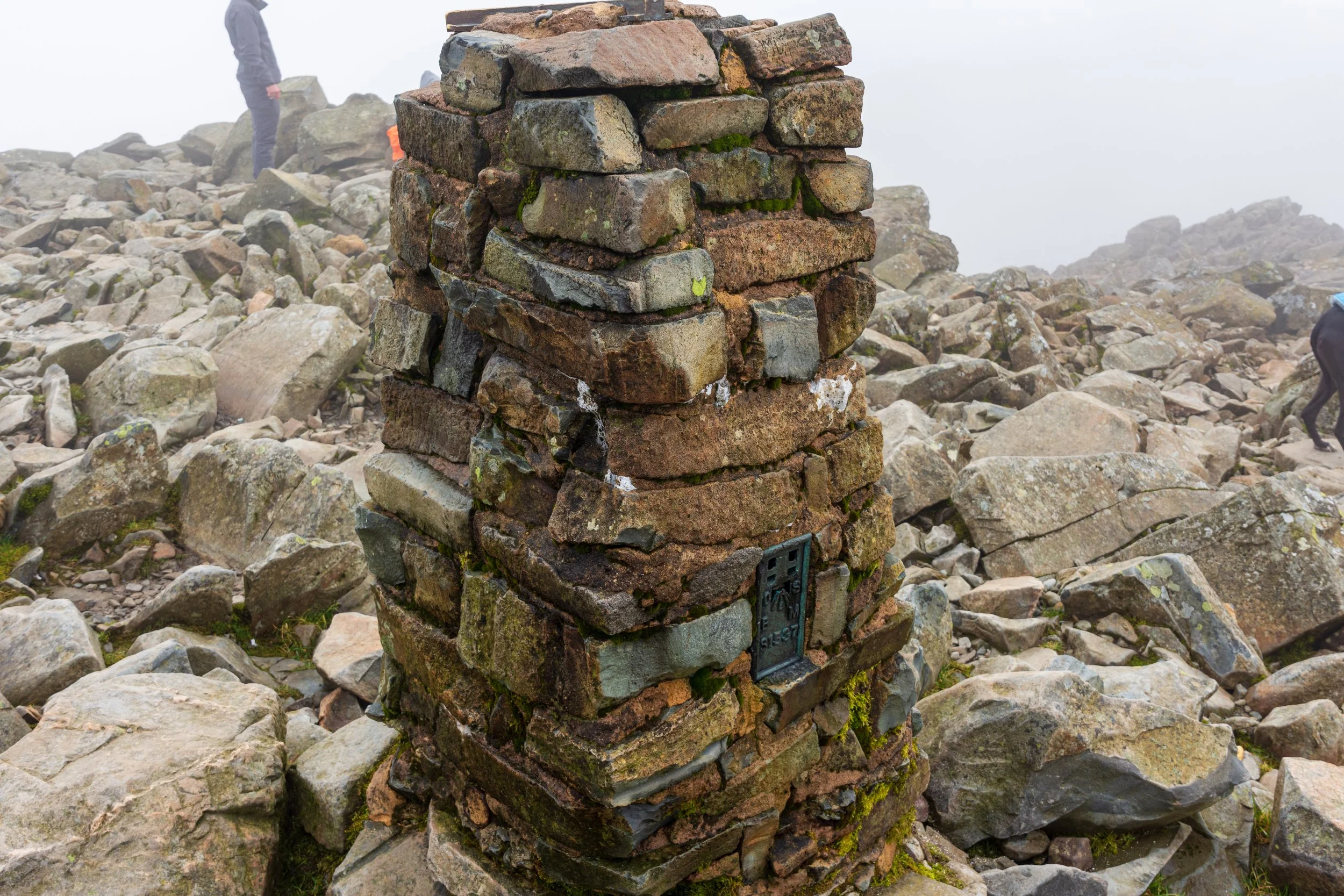 The brick-built triangulation pillar on the summit of Scafell Pike on a misty day, surrounded by a boulder field