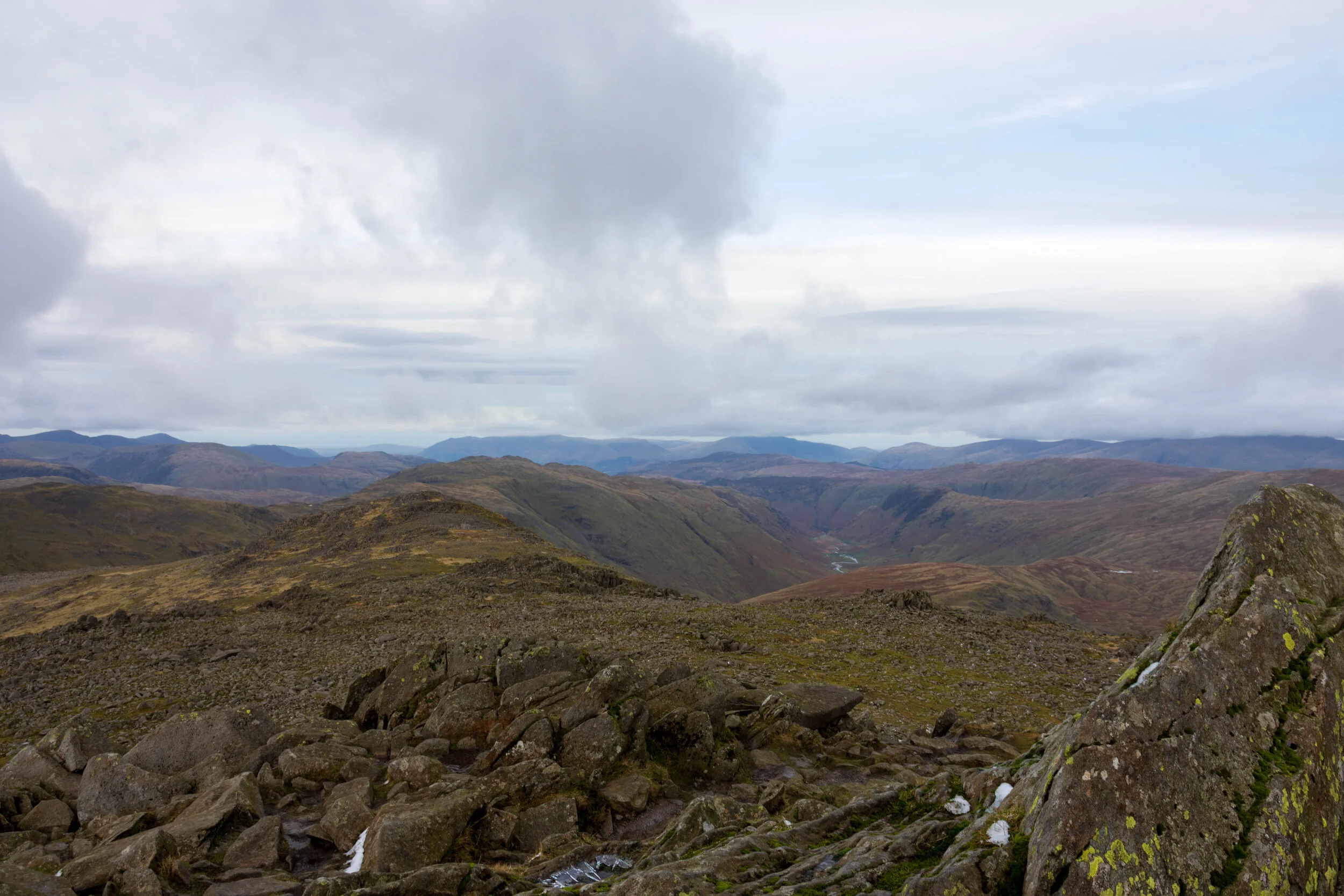 View from the rocky summit of Bow Fell in the Lake District, with mountains spreading far into the background, against a sky filled with light white clouds