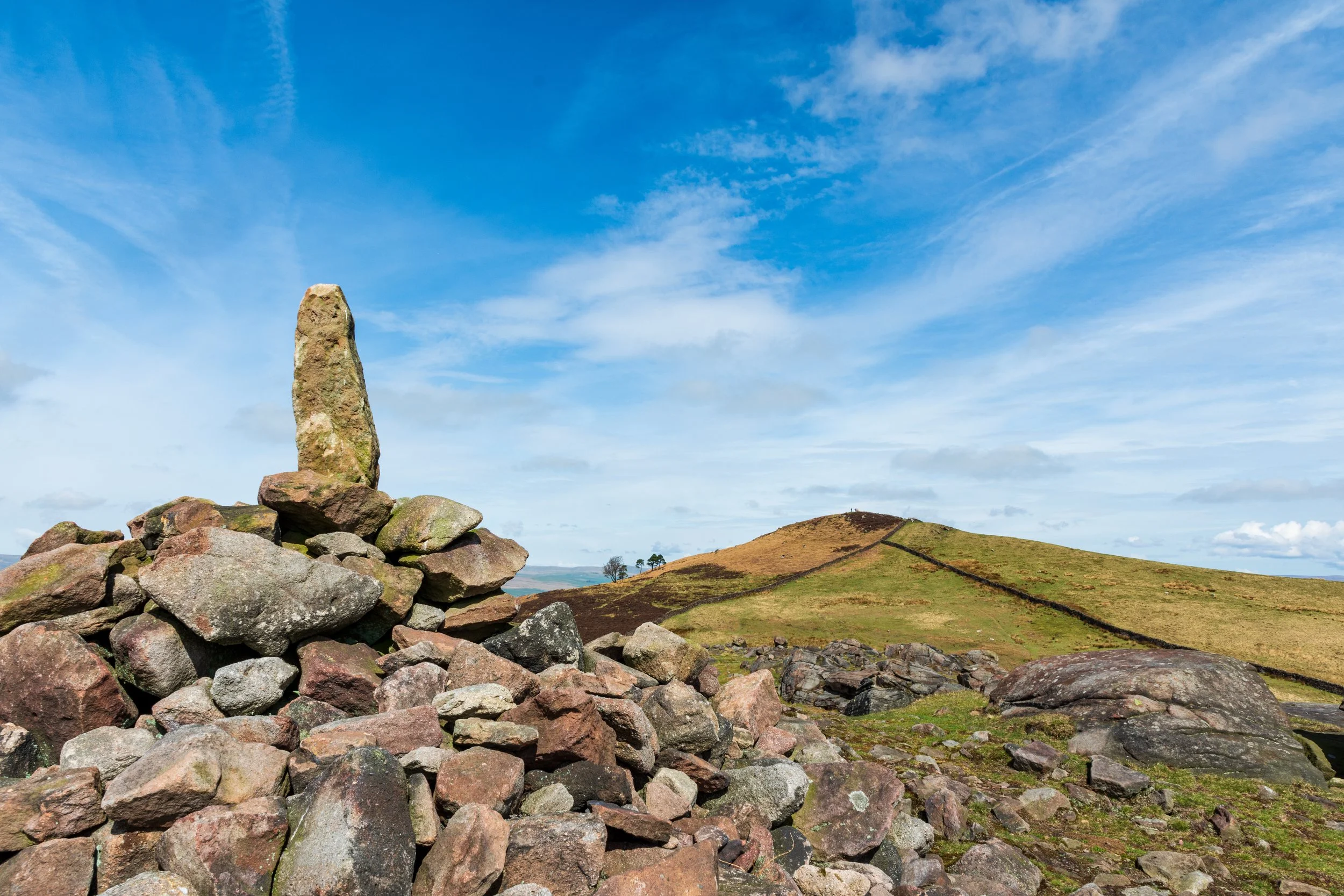 A summit stone, possibly ancient, placed on top of the cairn of an unnamed summit, with the Sharp Haw hill and summit in the distance, on a sunny day with blue sky