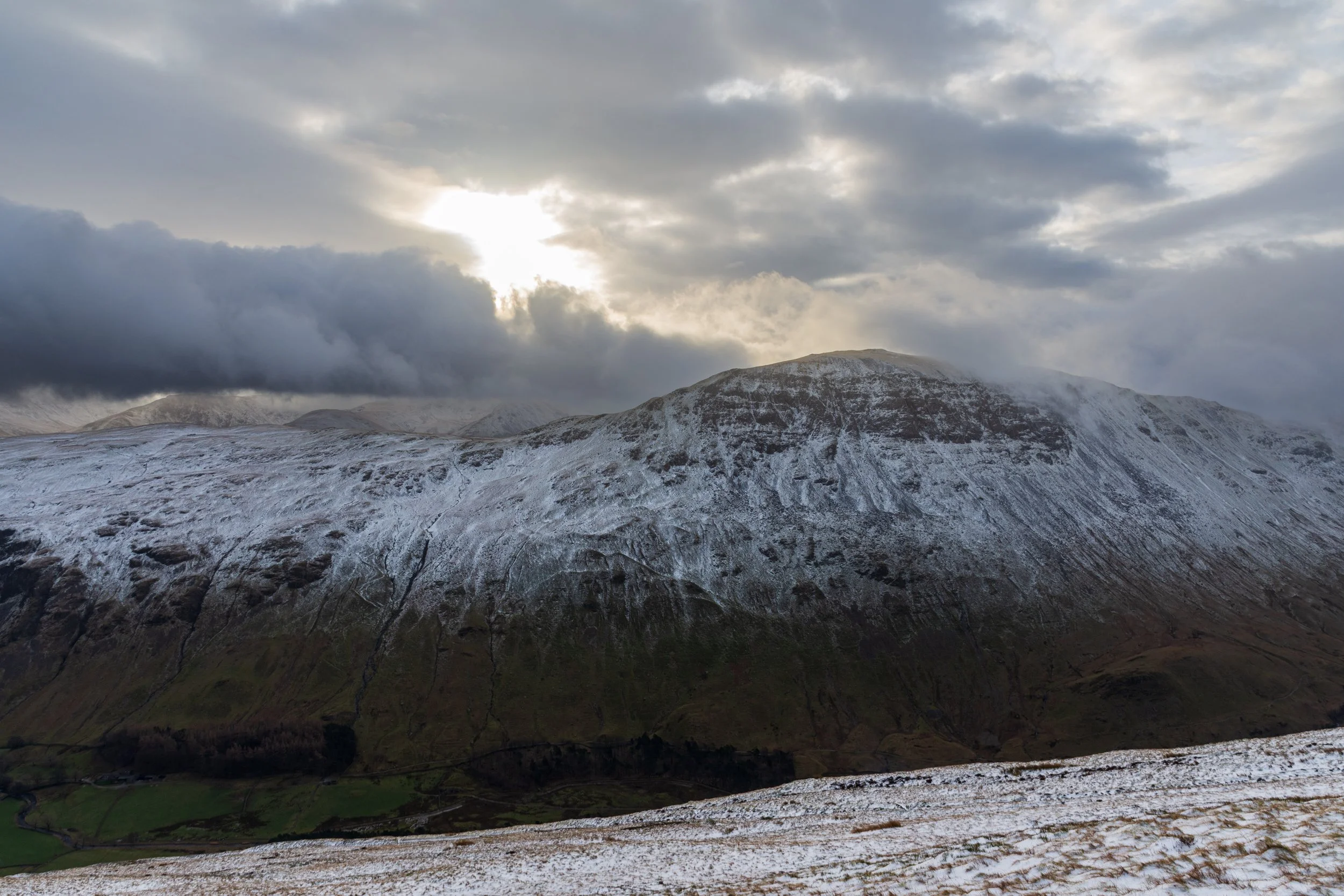 The snow-capped mountain of St Sunday Crag in the Lake District, viewed from Striding Edge, with low cloud hanging around the summits and the winter sun breaking through