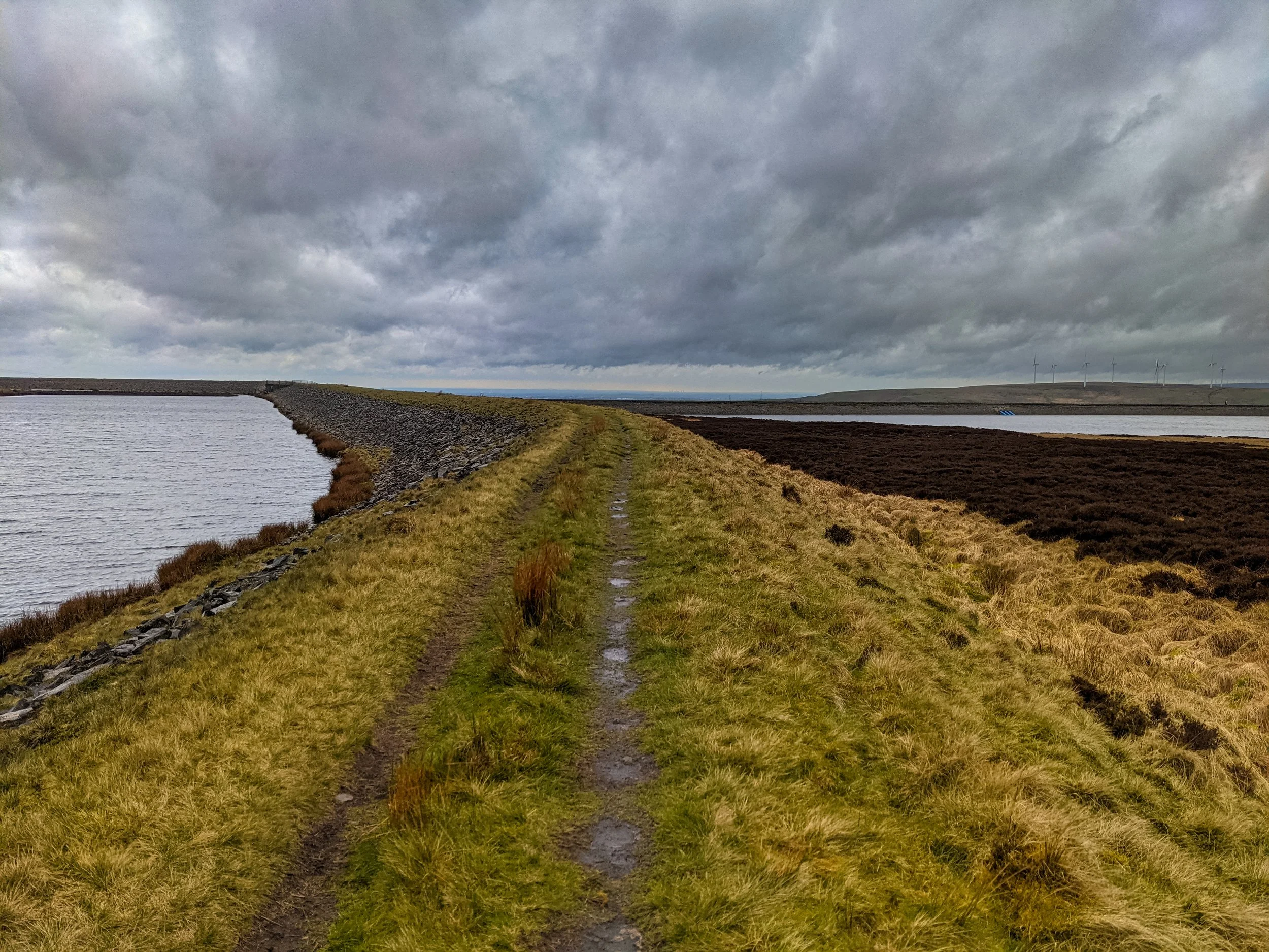A path between two reservoirs on the summit of Stoodley Pike, with wind turbines in the distance, and under a dramatic cloudy sky