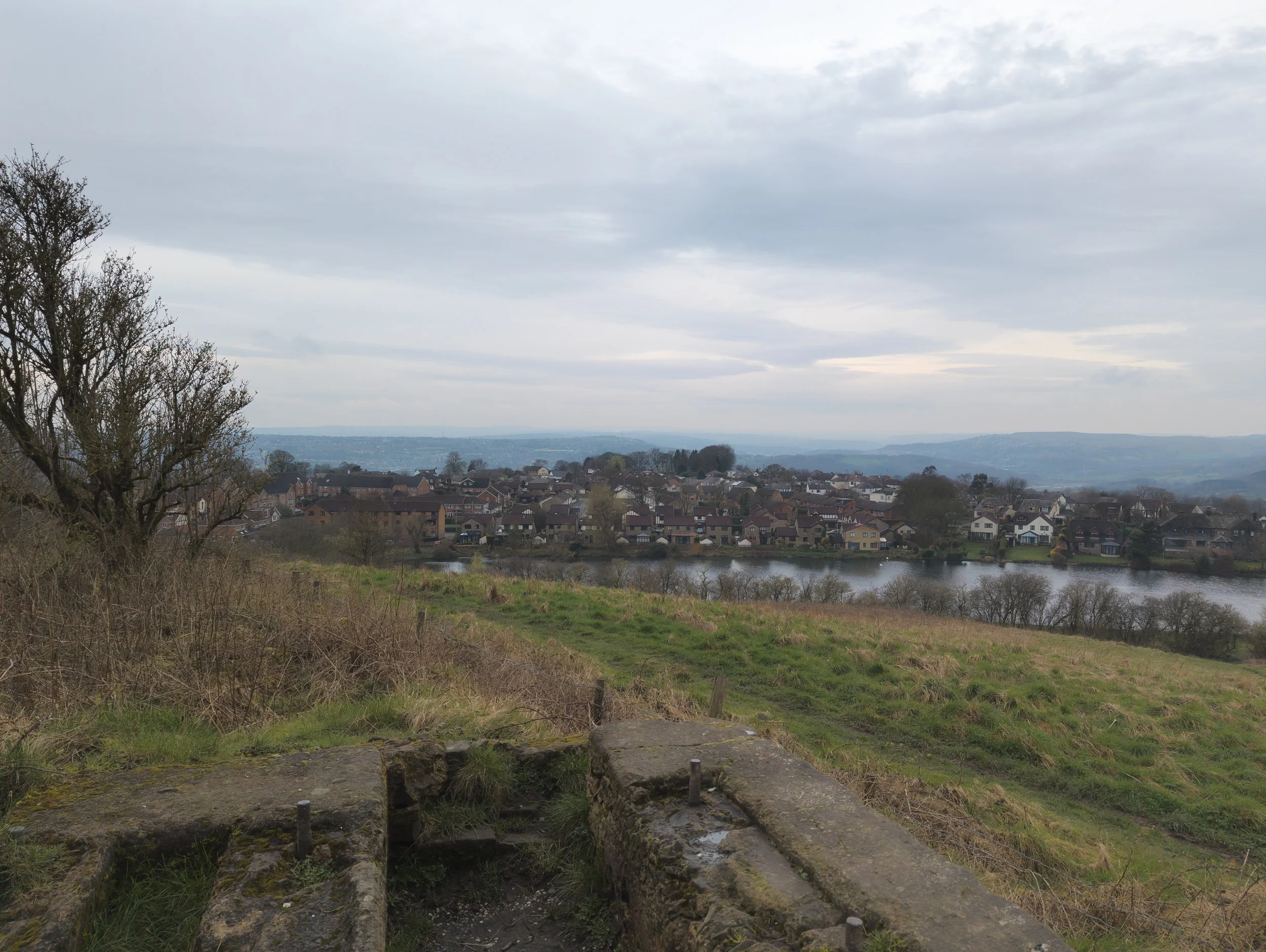 A view or Rawdon and Larkfield Dam from the slopes of The Rawdon Billing, with the West Pennine Moors in the background, concrete blocks in the foreground, on a cloudy day