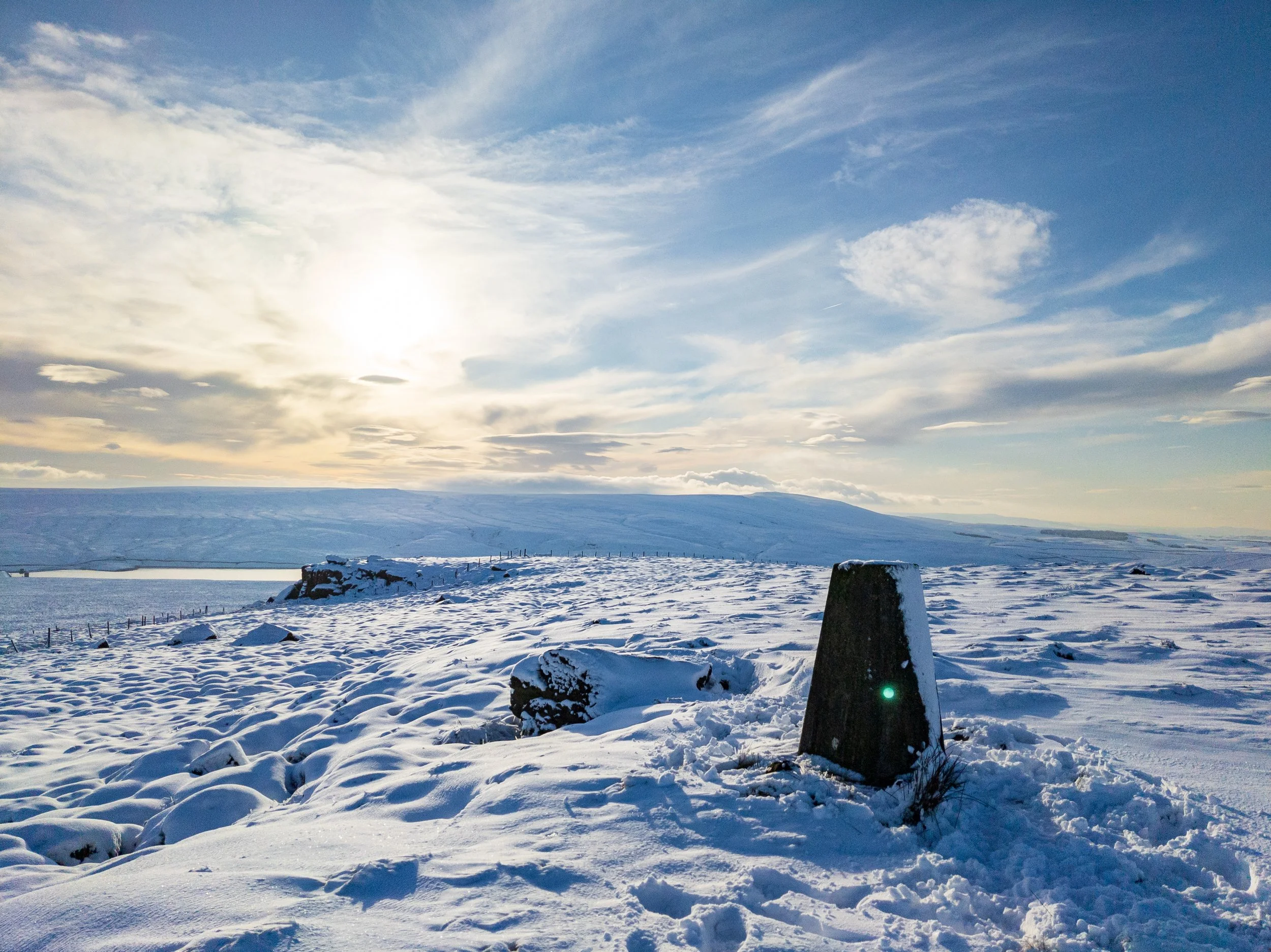 The trig point at Wolf Stones, on a snowy moor, on a sunny winter afternoon, looking towards Keighley Reservoir