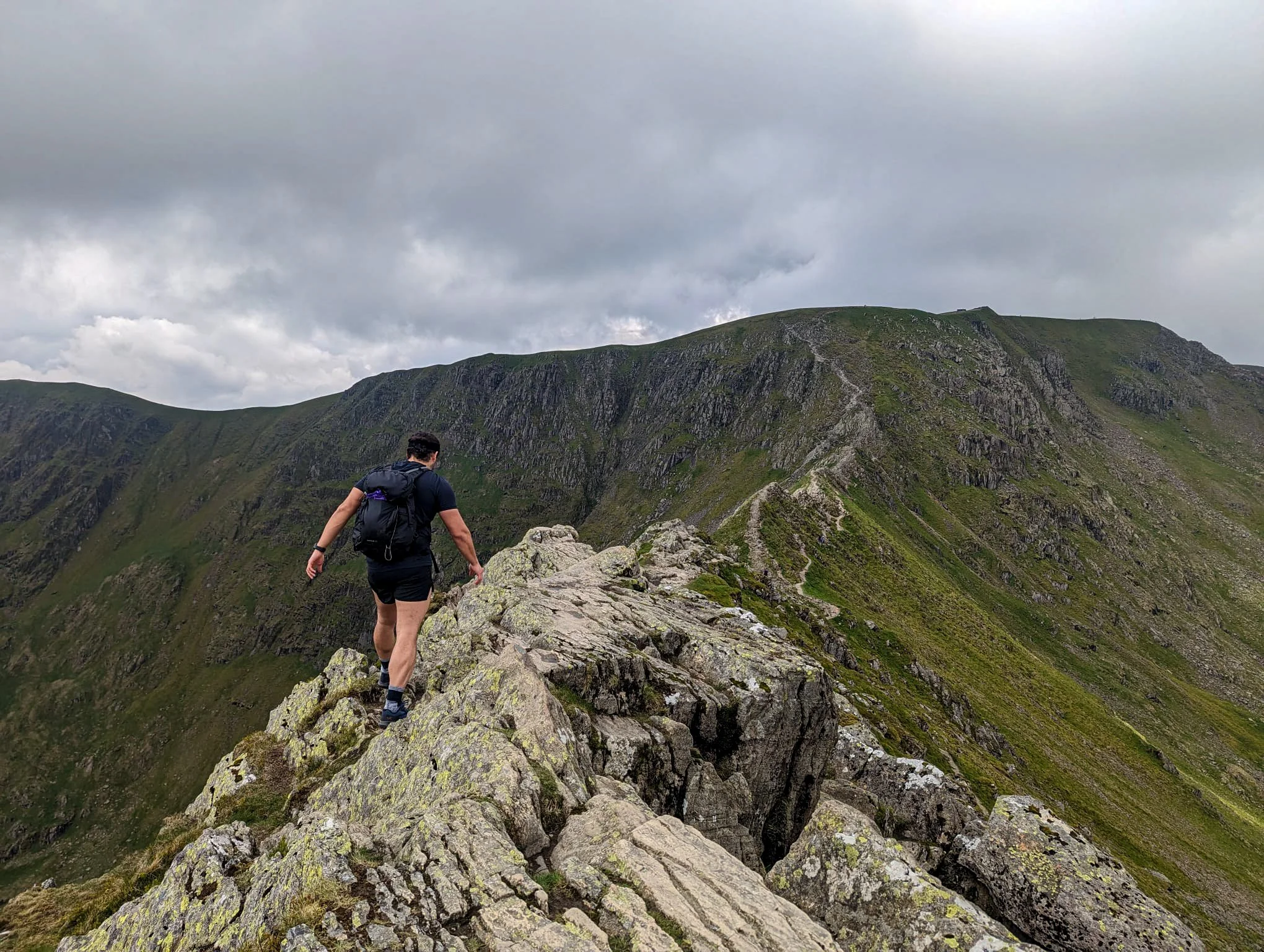 A man wearing black shorts and t-shirt, walking across the ridge of Striding Edge on the approach to Helvellyn on a cloudy, but clear day