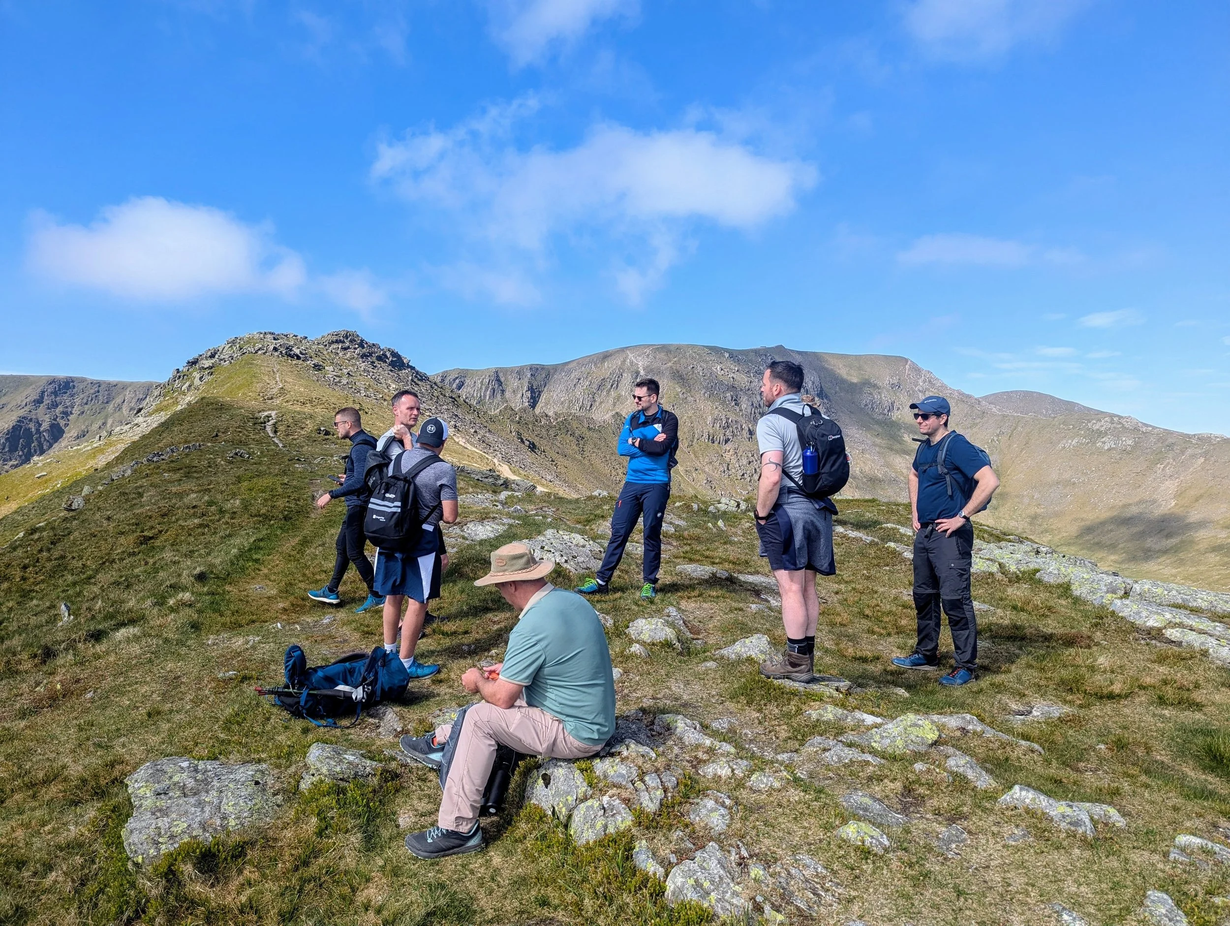 A corporate group of seven people on Striding Edge on a Mountain Boardroom Walkshop, wearing hiking gear, standing and sitting on a grassy hillside with rocky terrain, surrounded by mountains under a blue sky with some clouds.