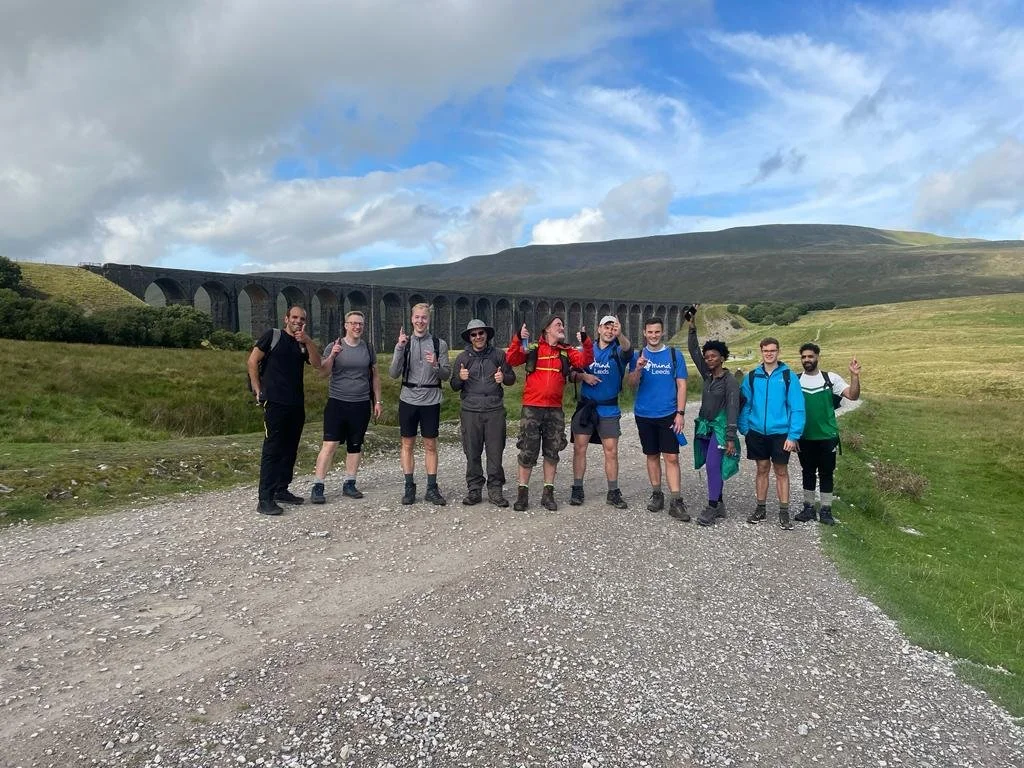 Group of hikers taking part in a Yorkshire Three Peaks Charity Challenge, standing on a dirt path in front of green fields and the stone Ribblehead viaduct, with hills (Whernside) and clouds in the background.