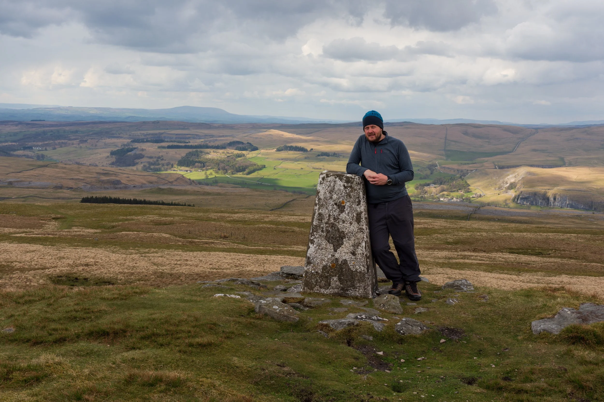 Ant Ackers, wearing outdoor gear with a Rab Fleece and North Face hat, leaning against a weathered trig pillar, with a view of Upper Wharfedale in the Yorkshire Dales, with Kilnsey Crag in the mid-ground and Pendle Hill in the background