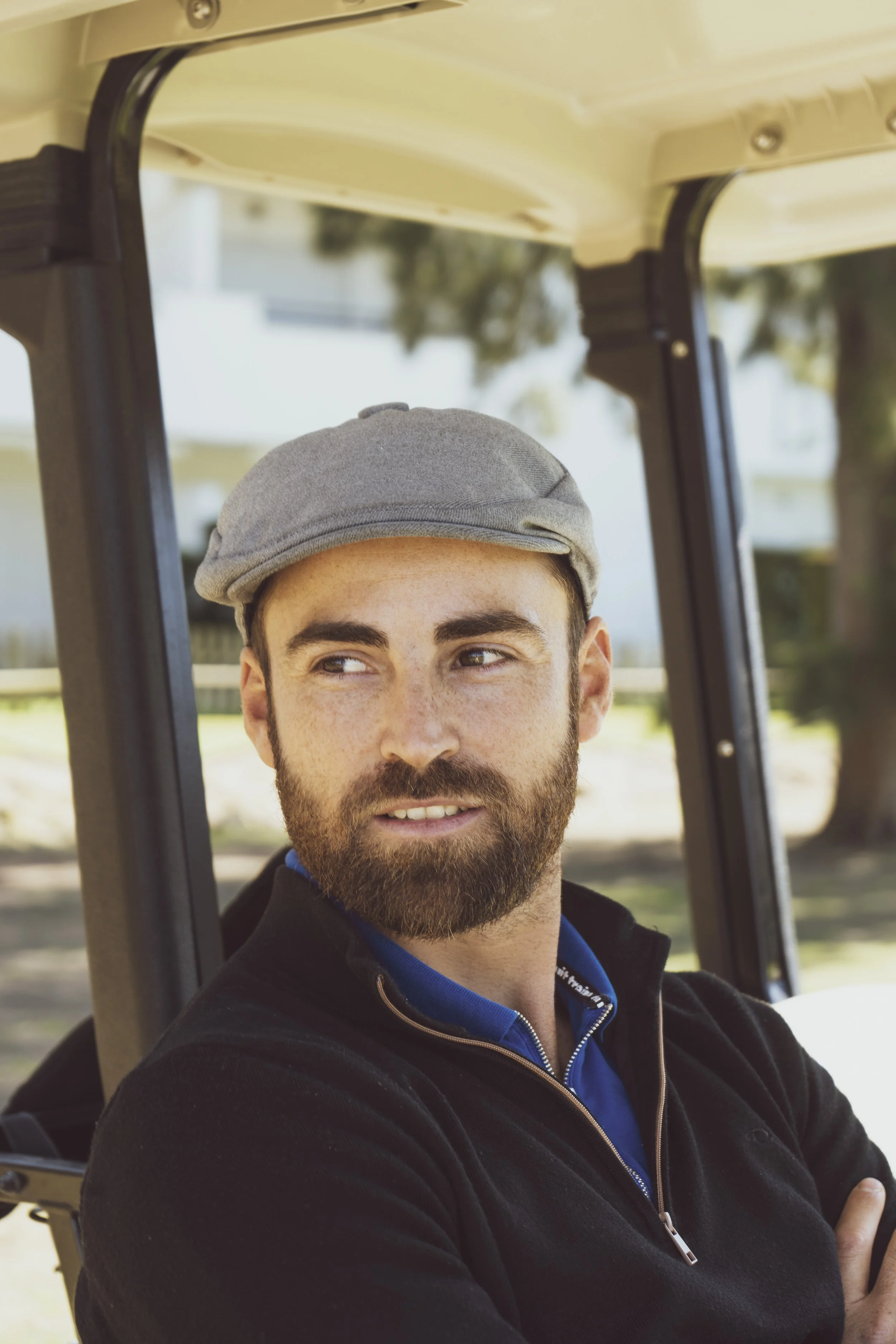 A man with a beard wearing a gray newsboy cap and a black jacket sitting in a golf cart.