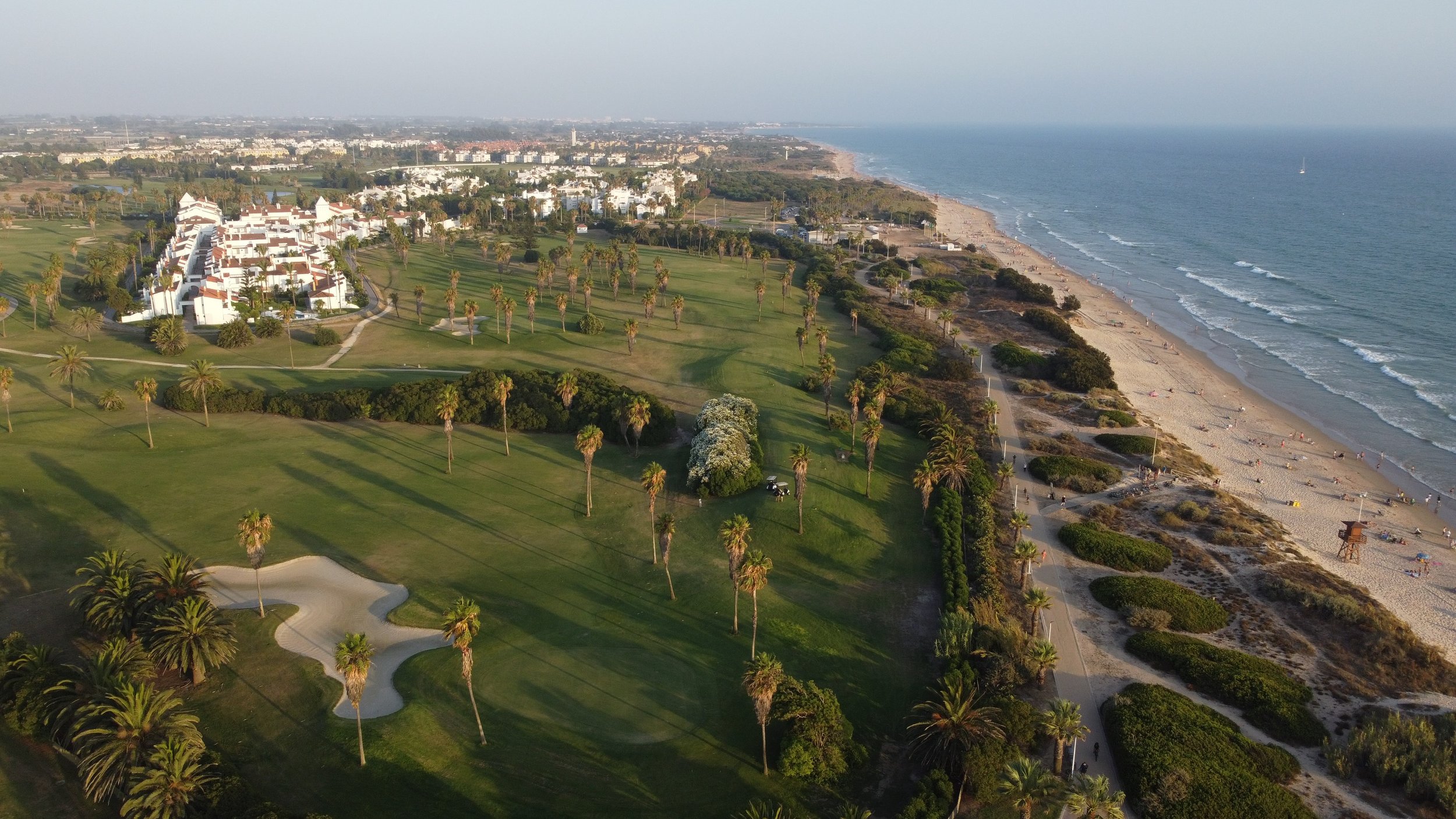 An aerial view of a coastal area featuring a golf course with palm trees, a beach with umbrellas and lounge chairs, and a cityscape in the background.