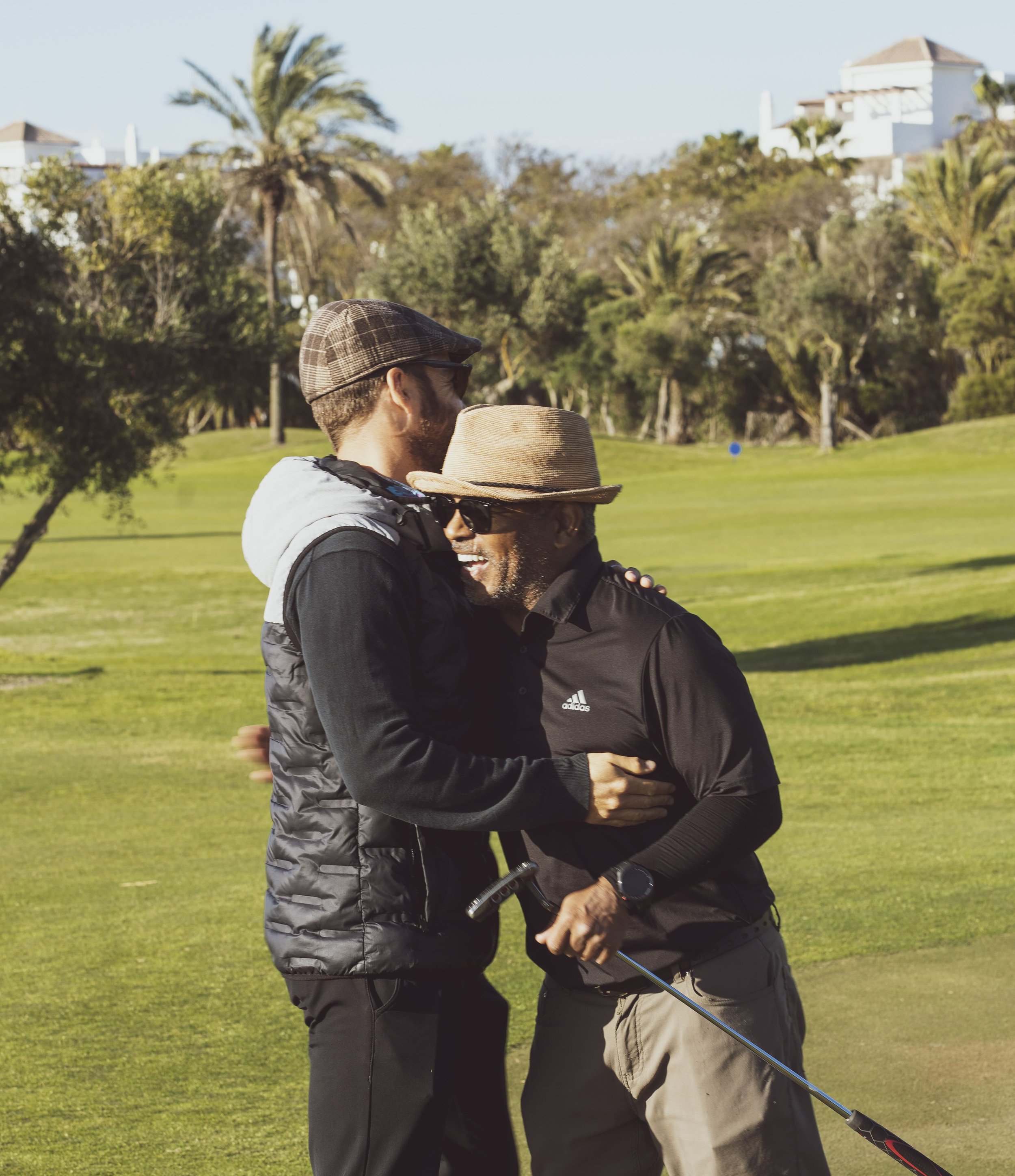 Two men hug on a golf course, smiling and holding a golf club.