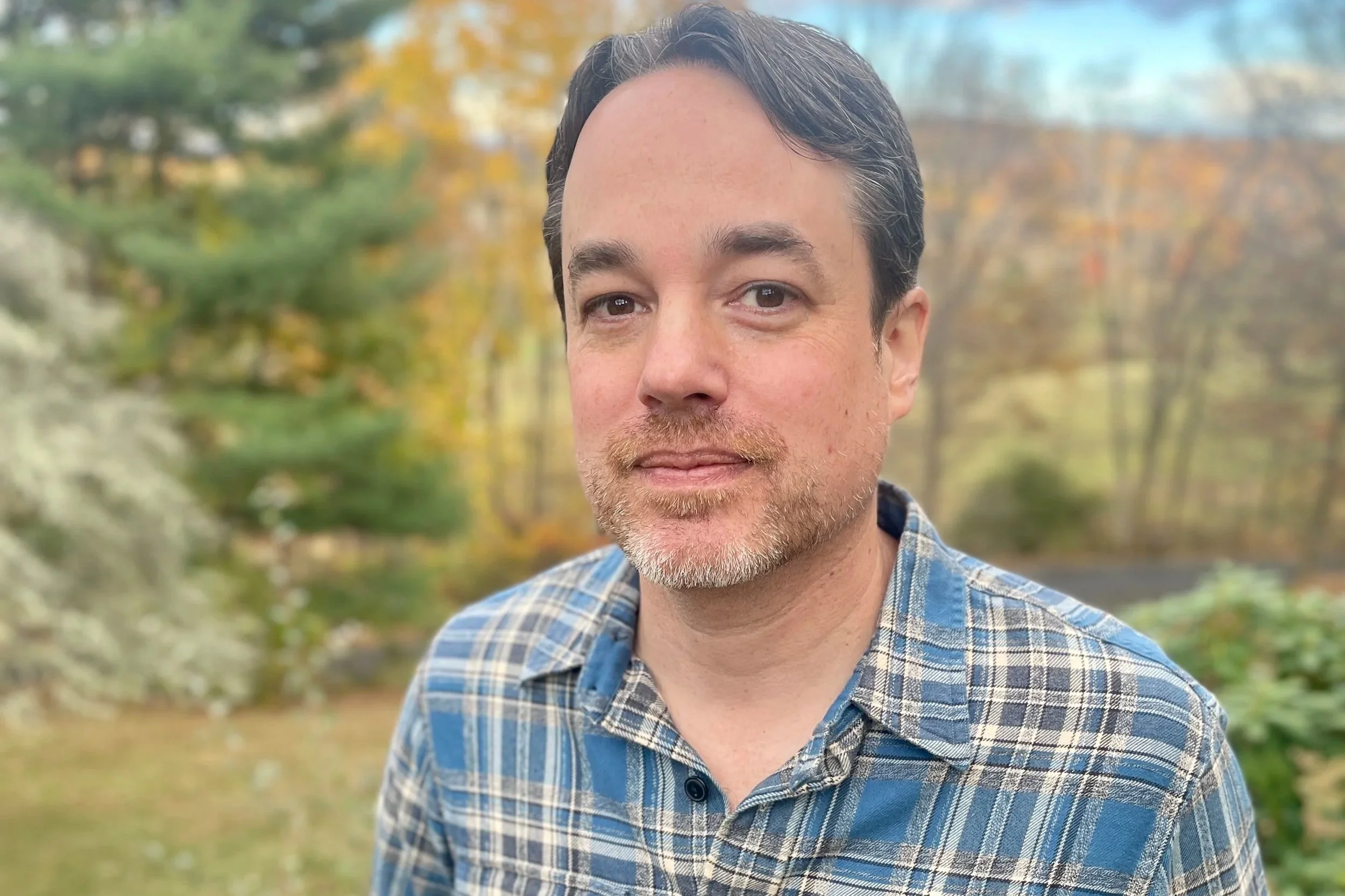 A man with dark hair and a beard wears a blue plaid button up shirt, leans back on a low wooden fence with autumnal trees and blue sky in the background.