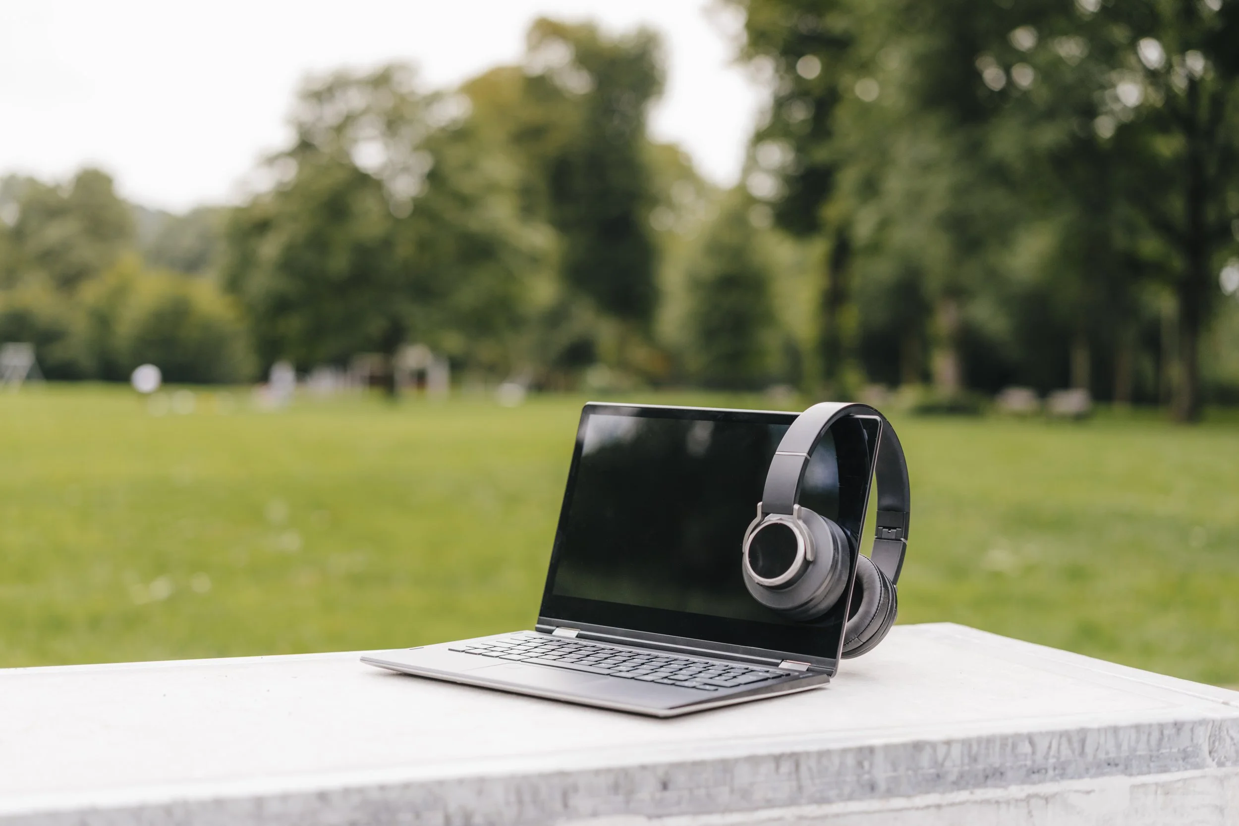 A laptop and headphones are on a table in a lush, green park.