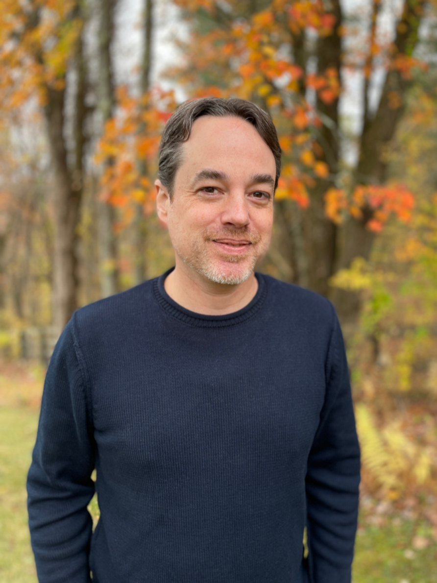 A man in a navy sweater smiles with autumn foliage in the background.