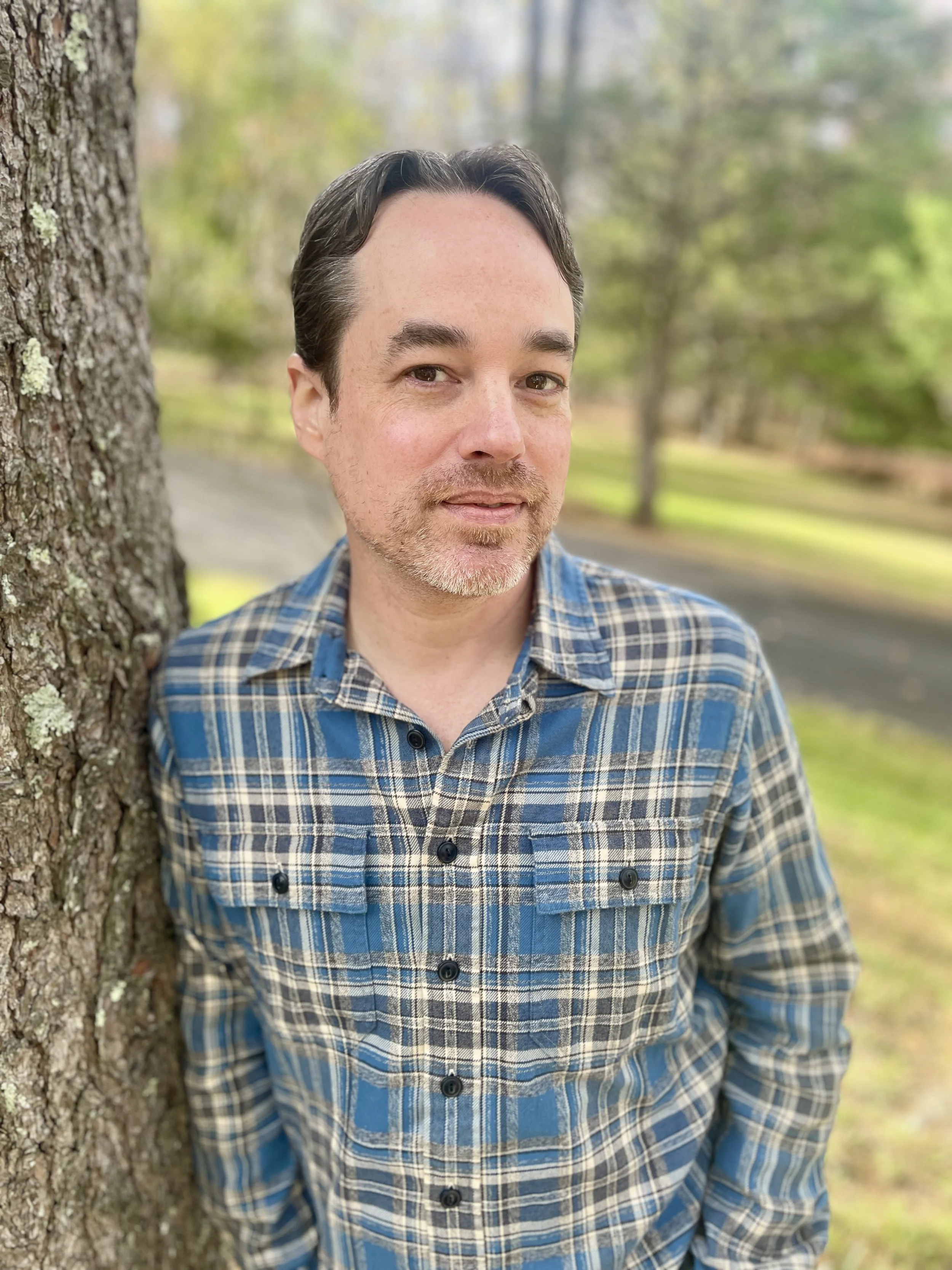 Man in a blue plaid shirt leans against a tree trunk with grass and trees in the background.