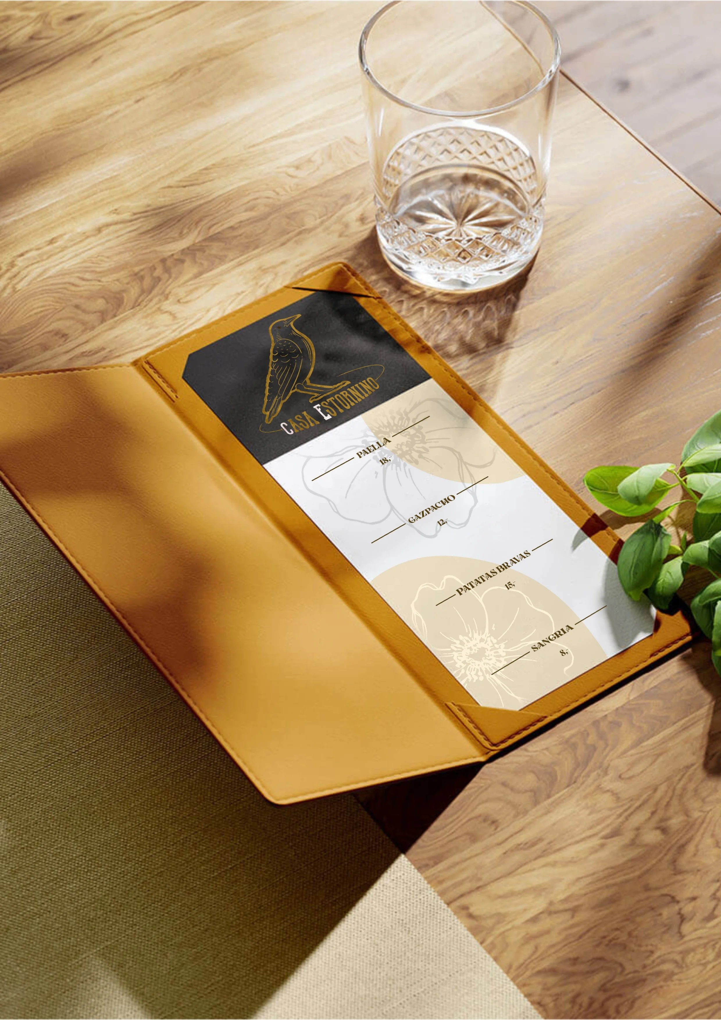 A restaurant menu inside a tan leather folder on a wooden table next to a glass of water and some green leaves.