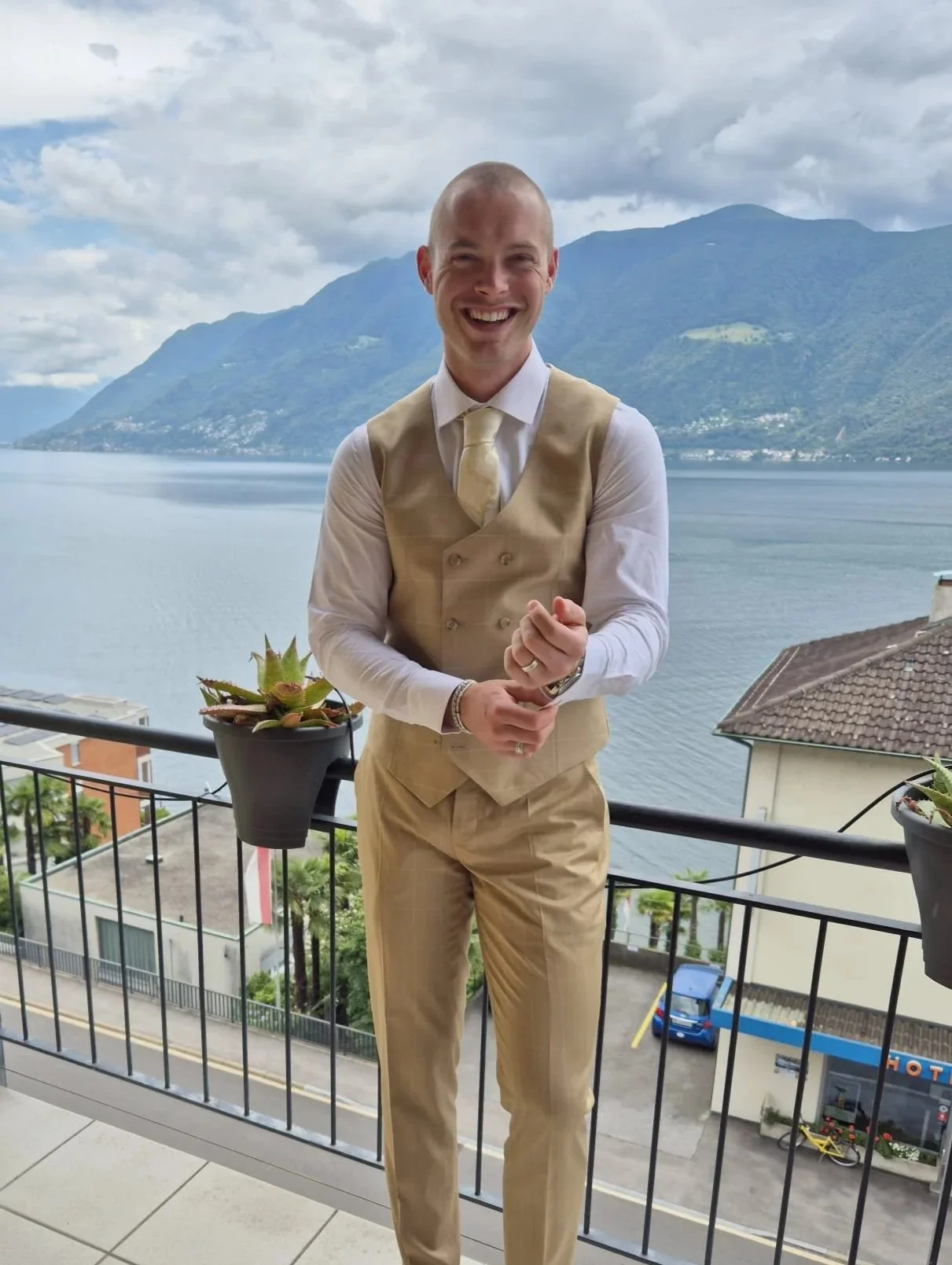 A man dressed in a beige vest, tie, and pants, smiling and standing on a balcony with a lake and mountains in the background.