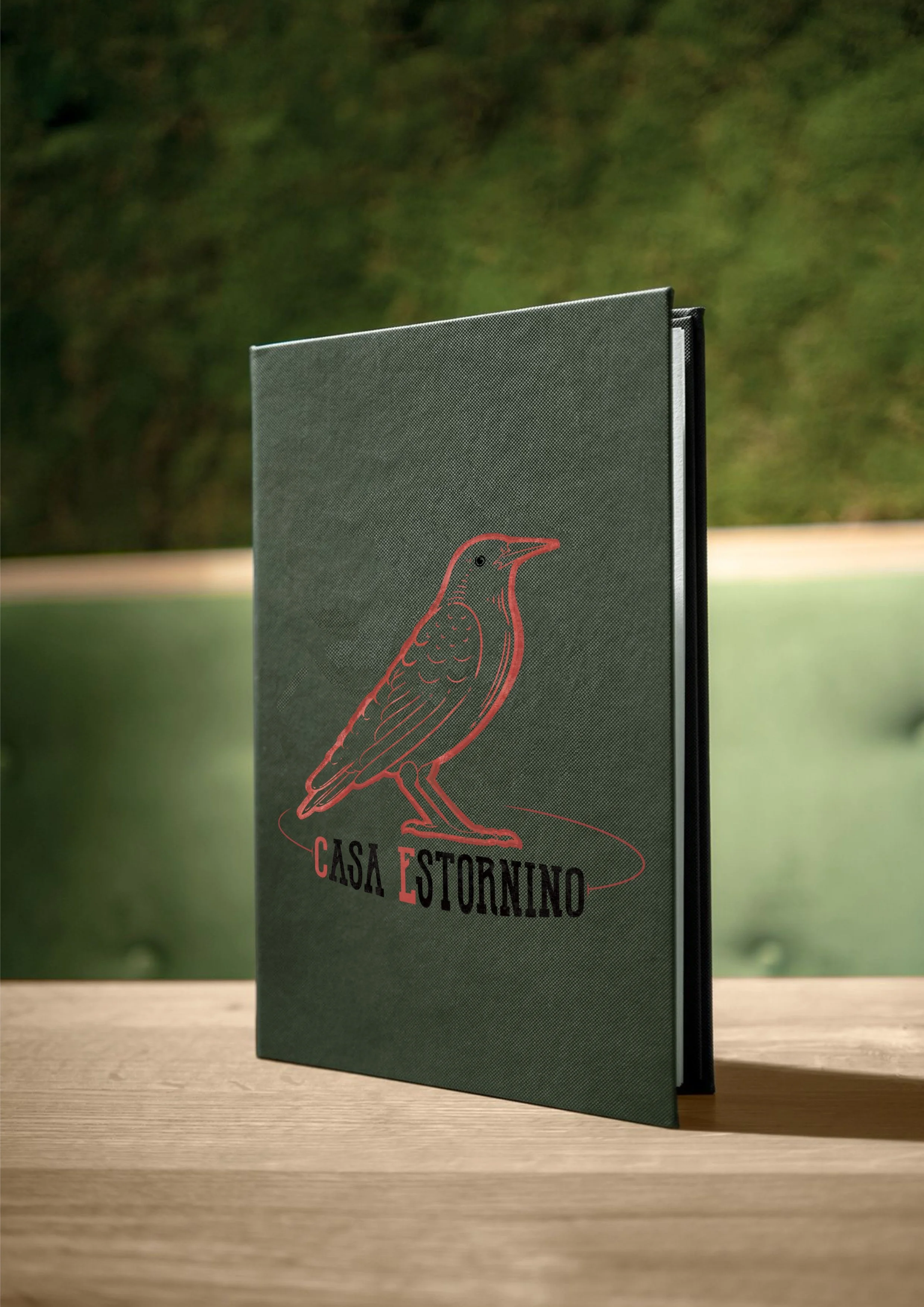 A menu cover with a bird illustration and the text "Casa Estornino" on it, placed on a wooden surface outdoors with greenery in the background.