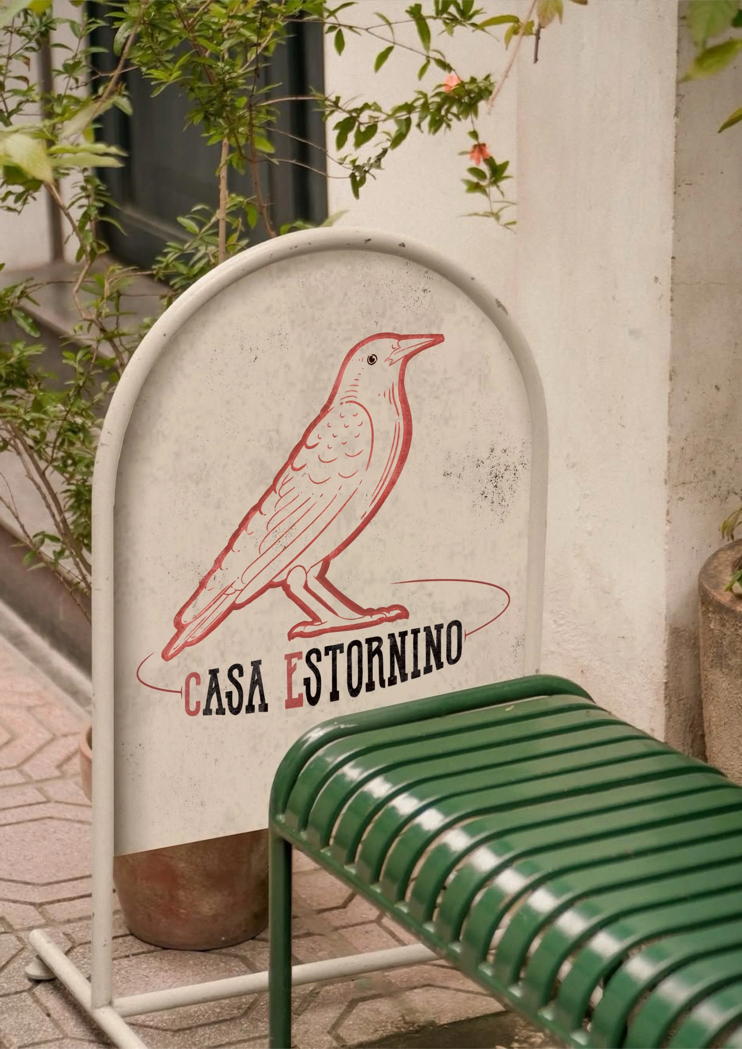 A white sign with an illustration of a bird and the text 'Casa Estornino', placed on the ground next to a green bench and some plants.