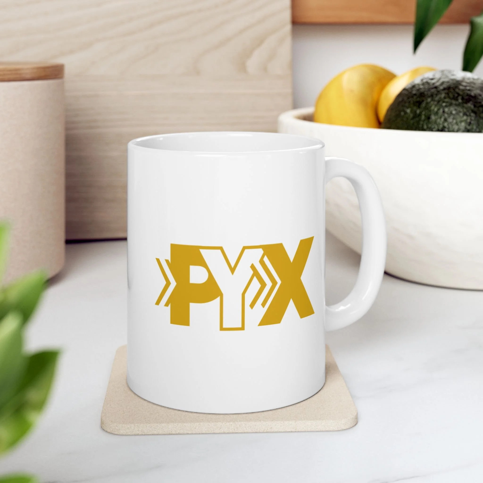 White coffee mug with yellow 'PYX' logo on a table with a beige coaster, with a bowl of fruit including yellow apples and avocados in the background.