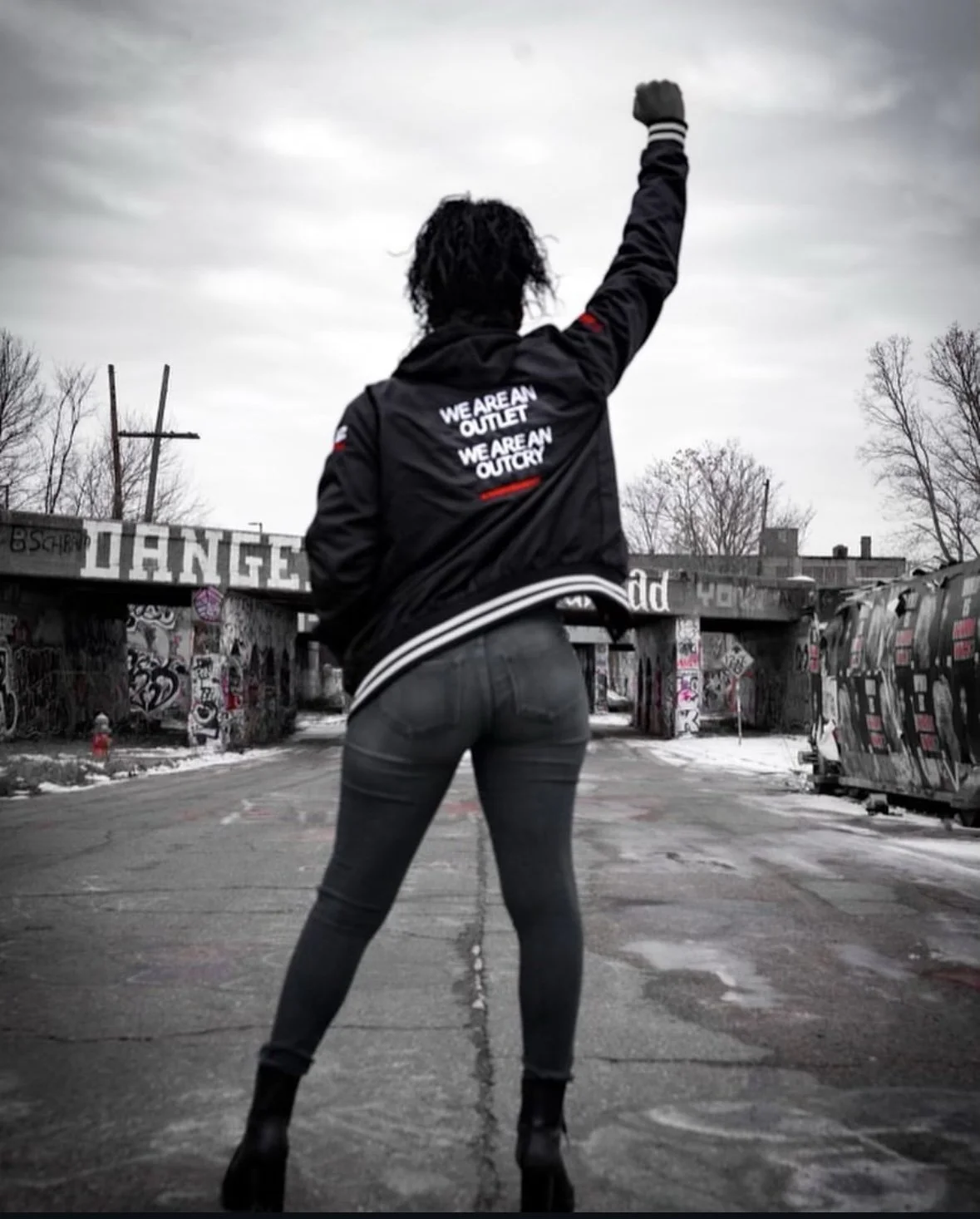 Back view of a woman with curly hair standing in an urban, graffiti-covered area with bare trees, wearing a black jacket with white text, dark jeans, and high-heeled shoes, raising her right arm in a fist.