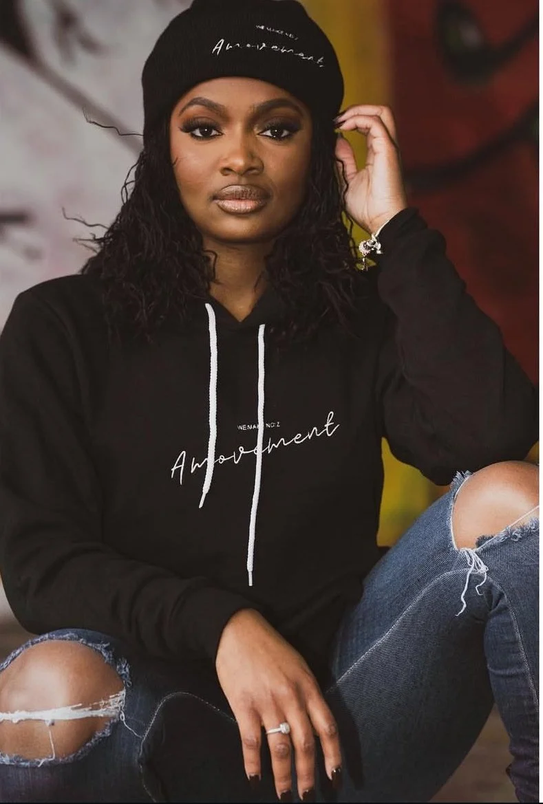 Young woman wearing a black beanie and black hoodie with white text, sitting with a relaxed pose, ripped jeans, and a bracelet.