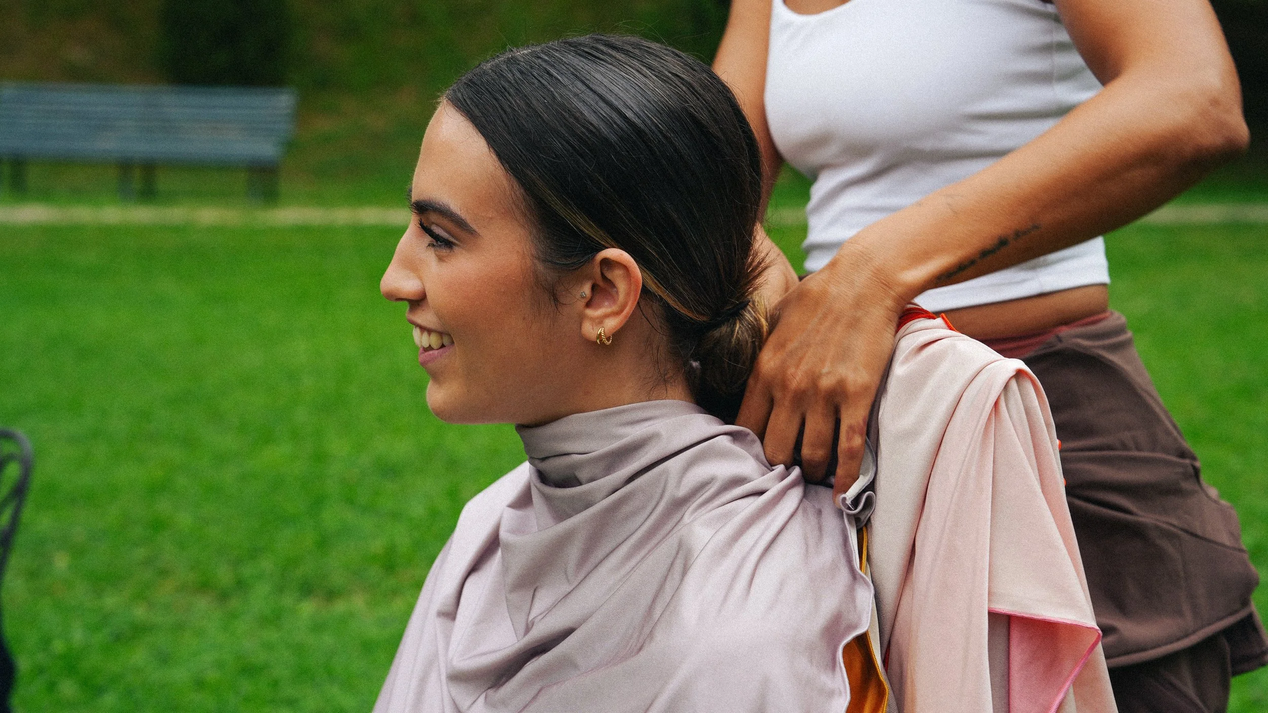Mujer sonriendo, recostada en un parque, con otra persona detrás haciendo ejercicios de estiramiento en el cuello.