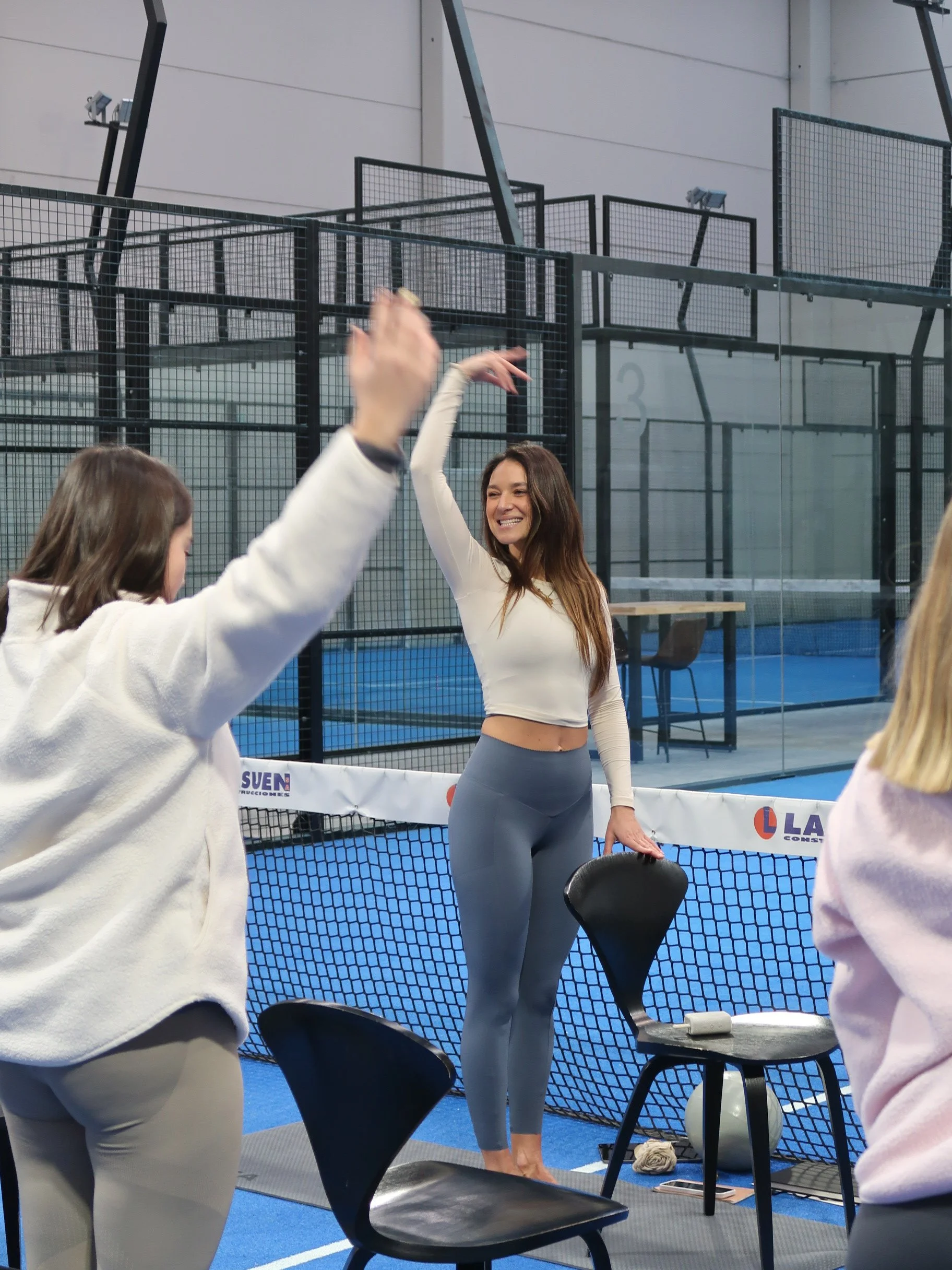 OHAZI EXPERIENCES: Mujer sonriendo y haciendo un gesto con la mano en un espacio deportivo cerrado, rodeada de otras personas, en lo que parece ser una clase o reunión deportiva en interiores.