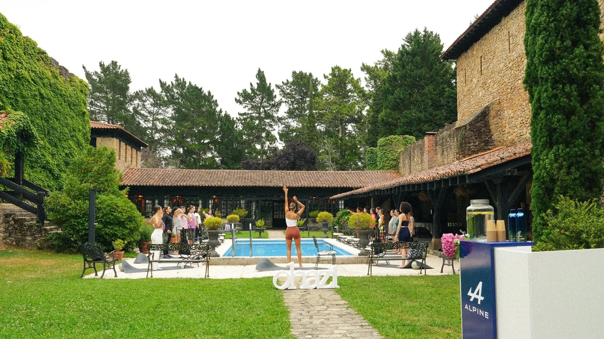 Grupo de personas realizando yoga junto a una piscina en un patio rodeado de árboles y edificios de piedra, durante el día.